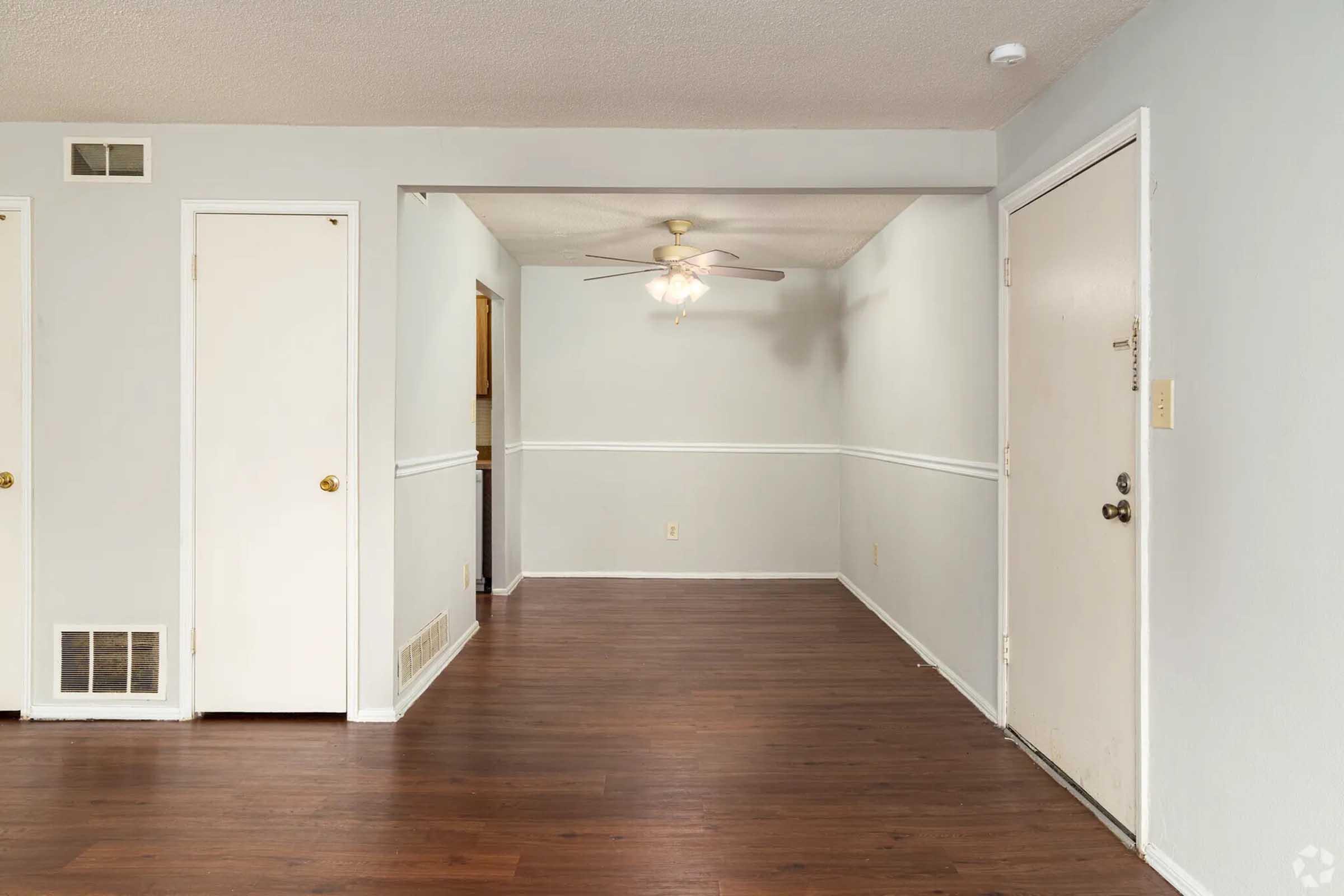 Interior view of a spacious room with light gray walls and hardwood flooring. A ceiling fan is visible, along with an entry door on the right and two closed doors on the left. The layout suggests a welcoming atmosphere, suitable for living or family spaces.