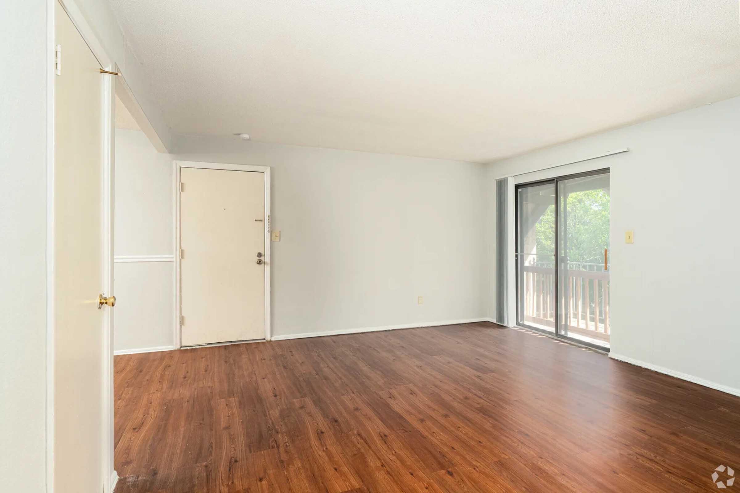 A spacious, unfurnished living room featuring light gray walls, a large sliding glass door leading to a balcony, and dark wood laminate flooring. The room includes a front door and a glimpse of natural light coming in from outside.