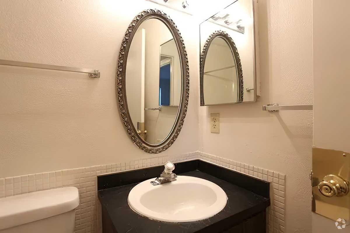 A small bathroom featuring a circular mirror with an ornate frame above a dark countertop and sink. The walls are light-colored, and there is a towel rack and lighting fixtures above the mirror. A toilet is visible to the left, and the overall design is simple and clean.