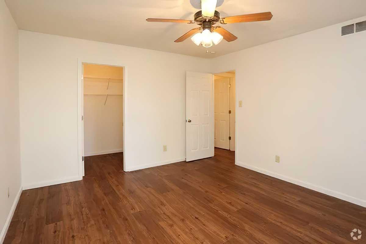A well-lit empty room with a ceiling fan, light fixture, and wood-style flooring. Two doors are visible: one leading to a closet and the other to another room. The walls are painted white, creating a spacious and clean appearance.