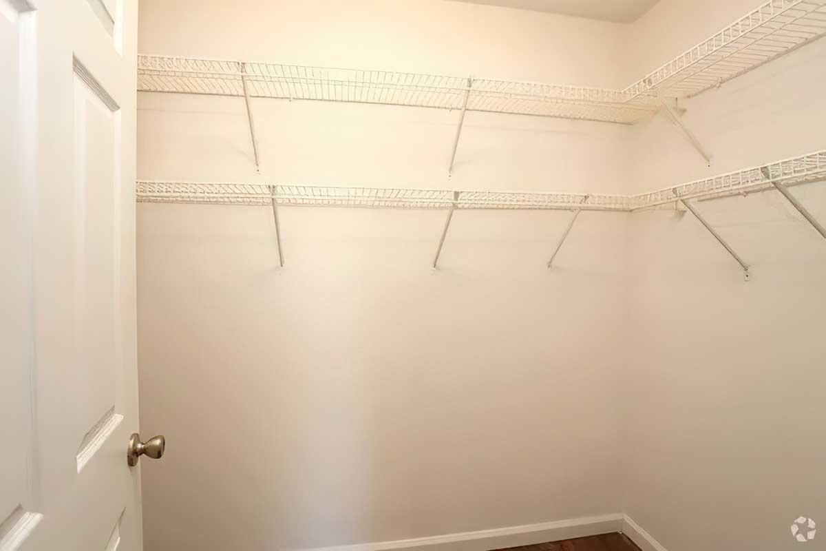 Empty walk-in closet with white wire shelving installed on the walls. The walls are painted a light color, and there is a closed door visible on the left. The floor has a wooden finish.