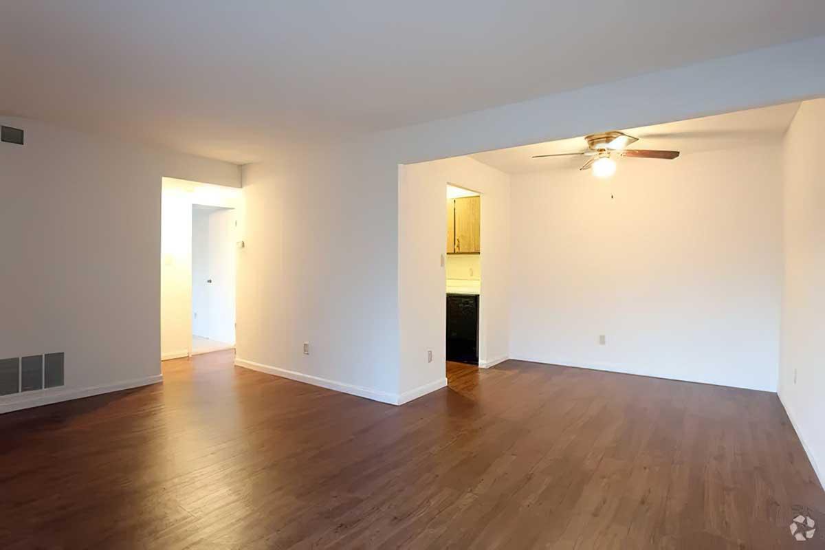 A spacious, empty room with hardwood floors, illuminated by a ceiling fan. To the left, a doorway leads to another room, while an open passageway connects to a kitchen area in the background. The walls are painted white, creating a bright and airy feel.