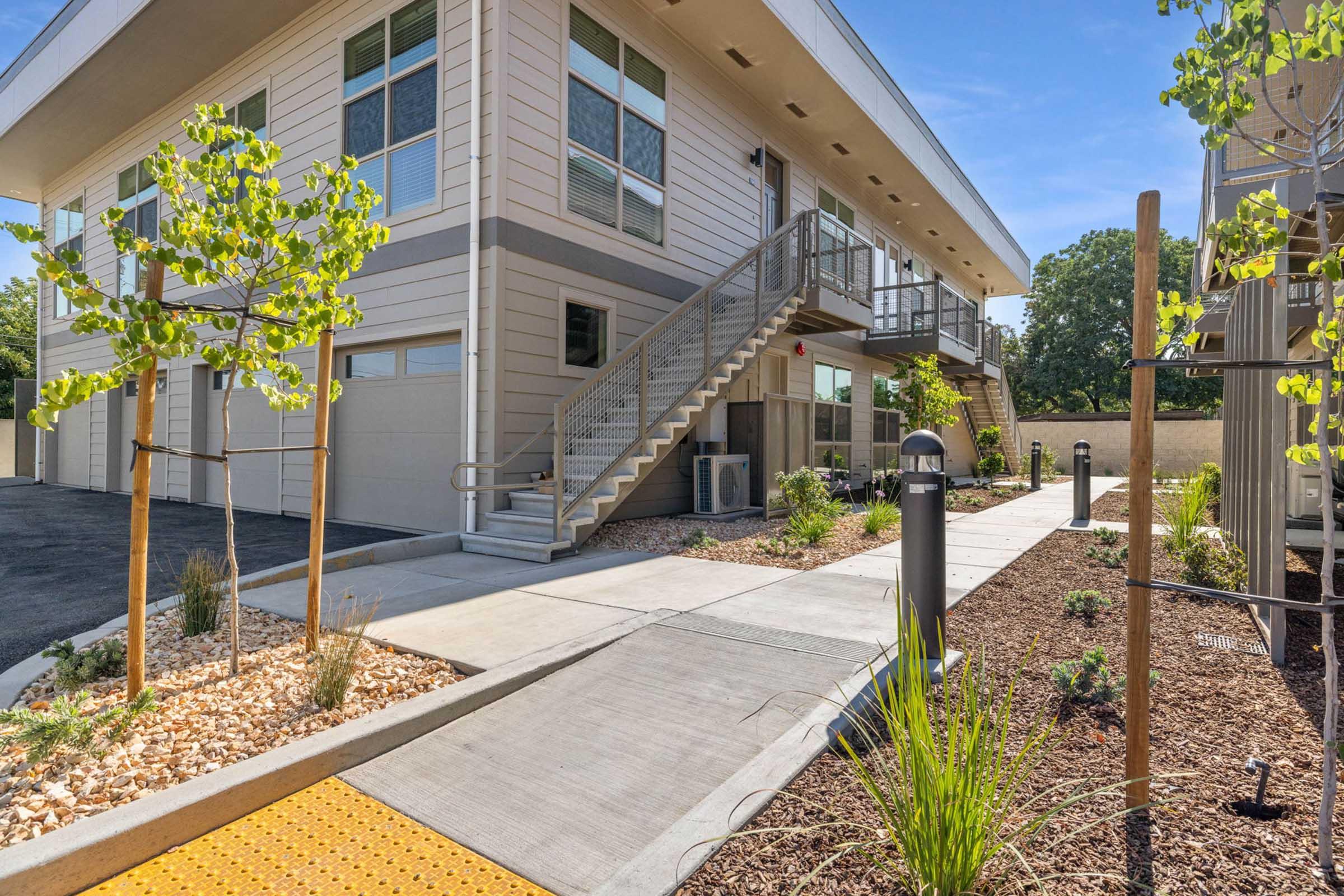 Modern two-story building with a landscaped entrance. The exterior features large windows and a staircase leading to the upper level. The pathway is lined with small plants and decorative gravel, while nearby rounded light posts add an inviting touch to the setting.