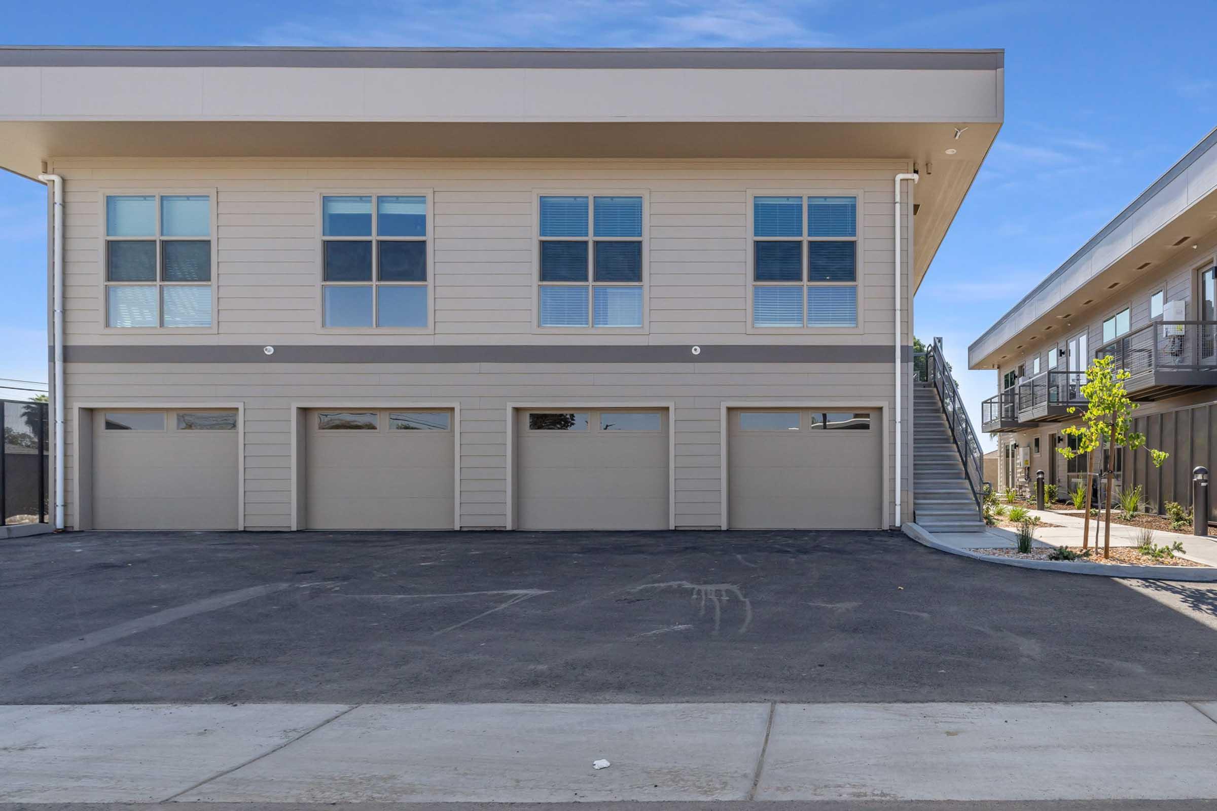 Two-story building featuring a modern design with multiple windows on the upper floor. The ground level has three garage doors, and a staircase leads to the second floor. The surrounding area is paved, with some landscaping visible on the right. The sky is clear, indicating a sunny day.