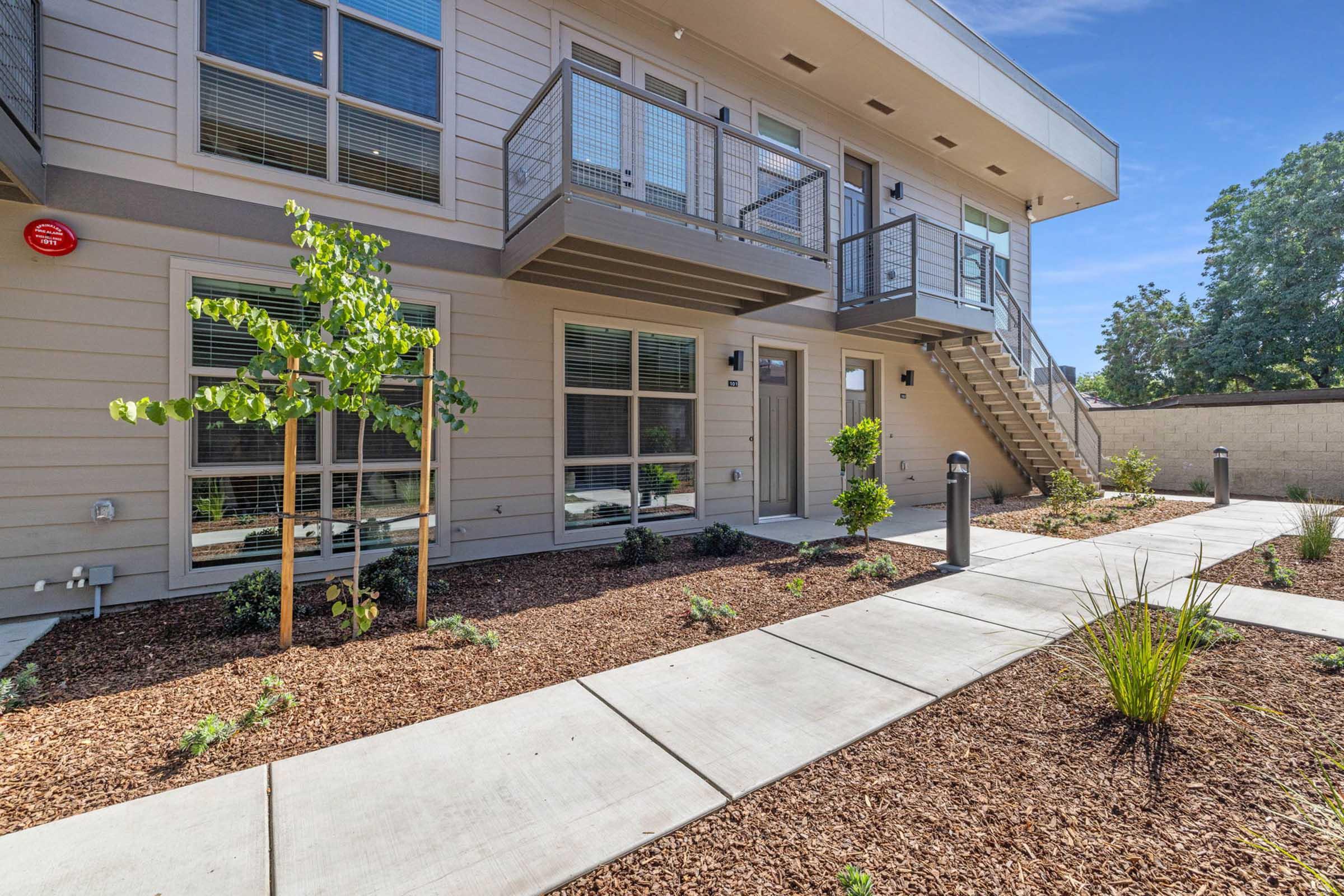 View of a modern, multi-unit residential building with a landscaped courtyard. Features include a concrete pathway, shrubs, and small trees. The building has multiple balconies, large windows, and a staircase leading to the upper floor. Bright blue sky in the background adds to the inviting atmosphere.