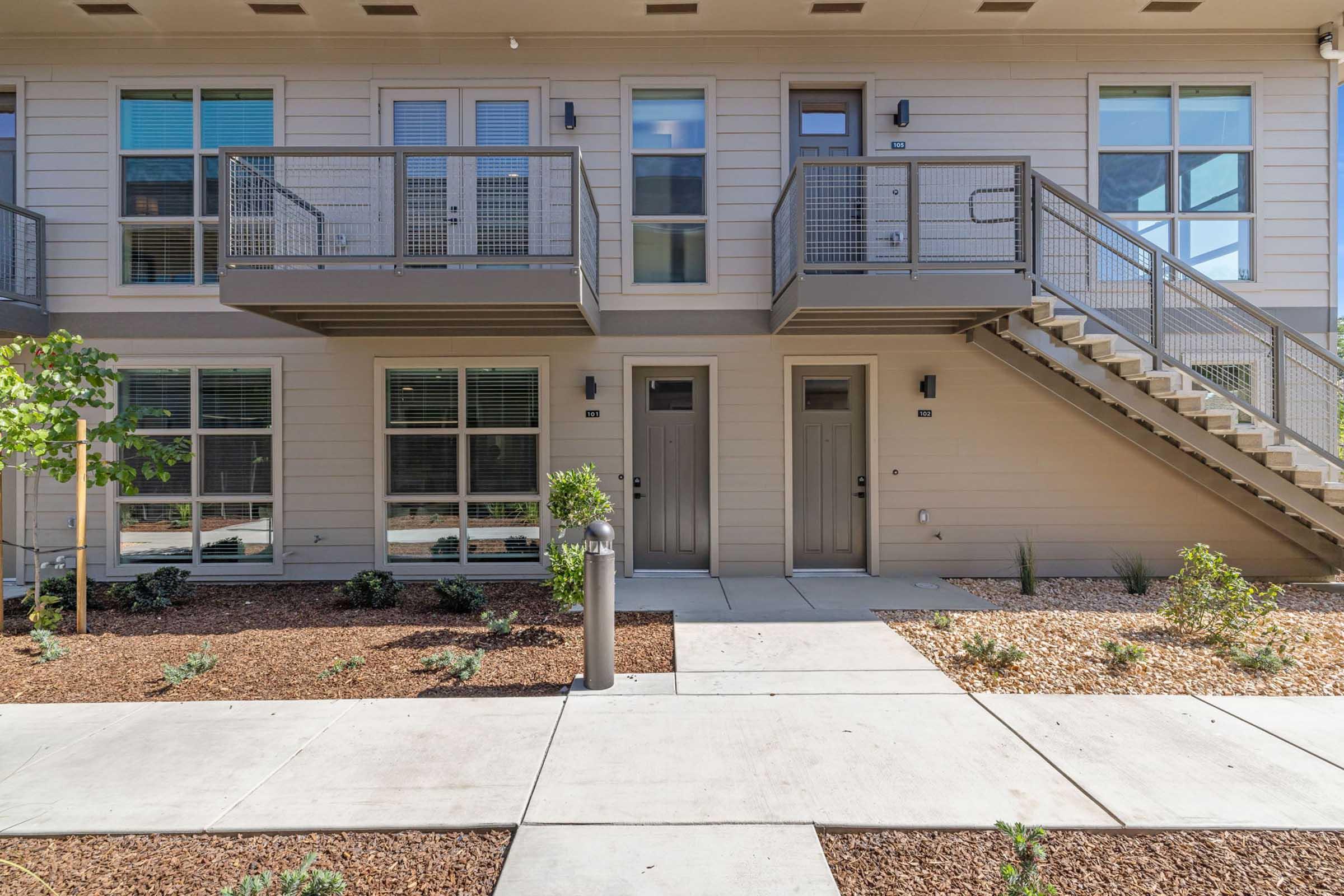 A modern apartment building features two levels with multiple entrances. Each unit has a small patio space. The exterior is light-colored with large windows, and there's a staircase leading to the upper level. Landscaping includes small shrubs and gravel patches along the pathways.