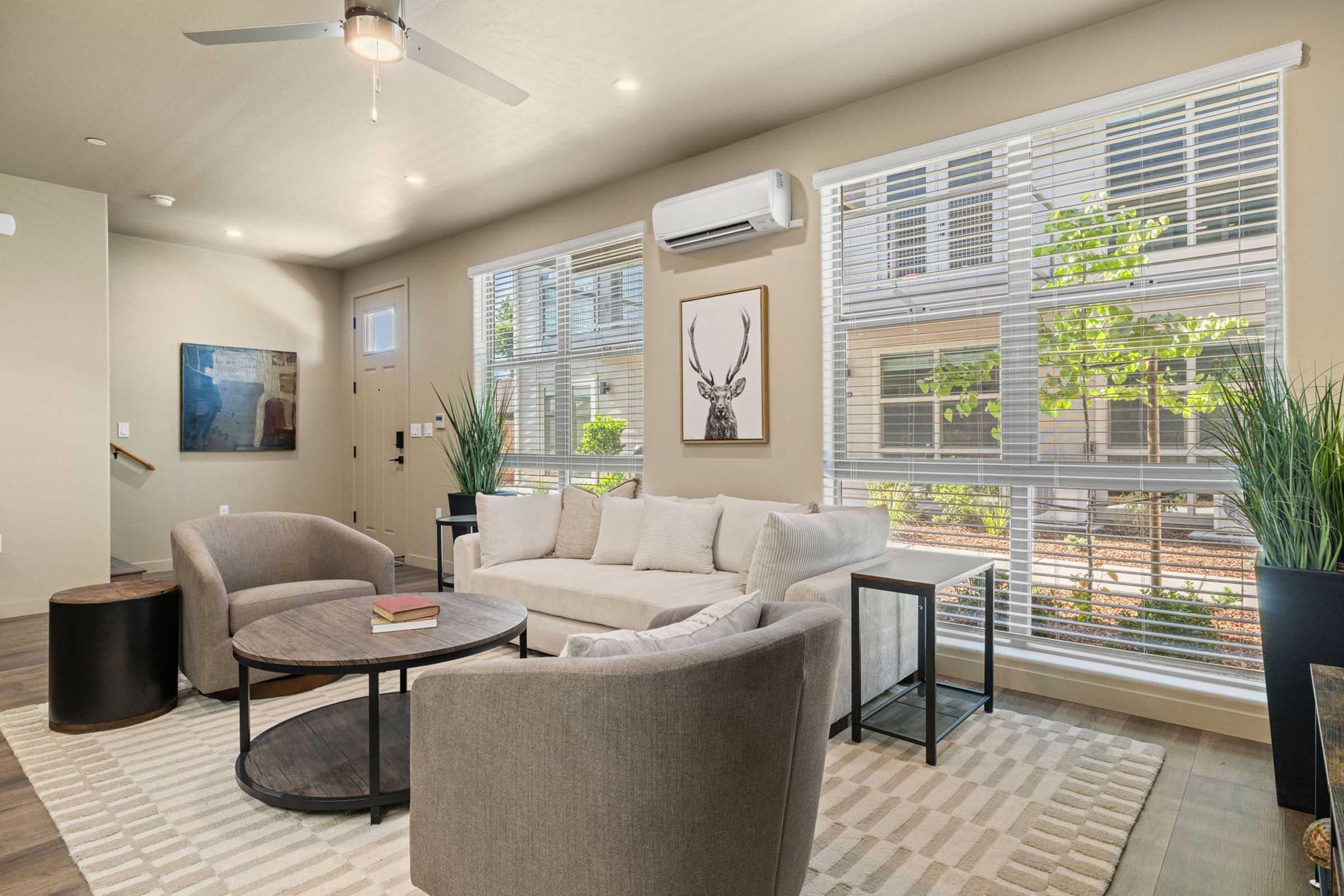 A modern living room featuring a light-colored sectional sofa, two round coffee tables, and stylish accent chairs. Large windows with blinds provide natural light and a view of greenery outside. A framed artwork of a deer adds decorative charm to the space, complemented by indoor plants.