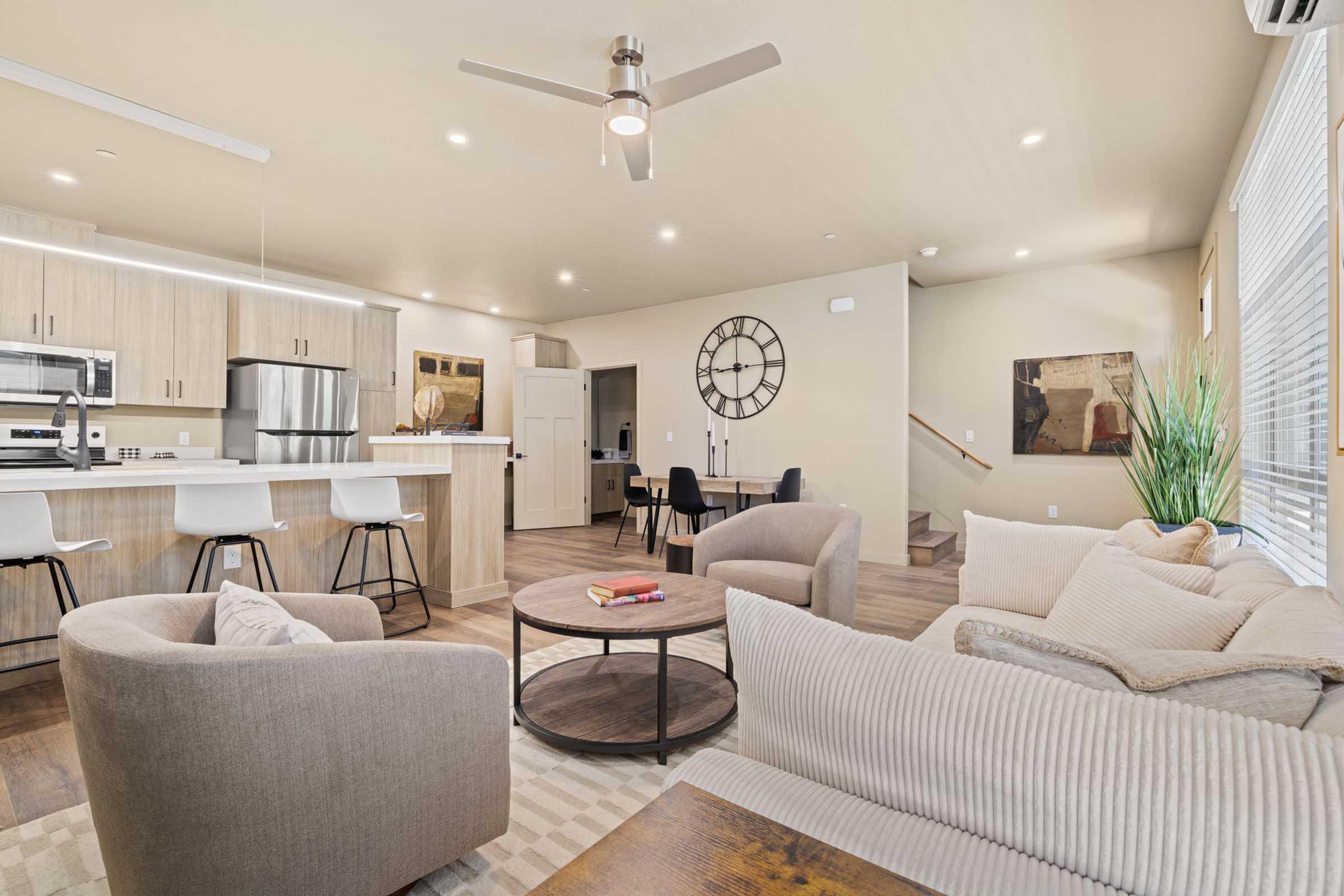 A modern, stylish living area featuring a comfortable beige sofa, a round coffee table, and a ceiling fan. The kitchen is visible in the background with wood cabinetry and stainless steel appliances. A large wall clock adds character, and there are plants and artwork to enhance the decor. Light-colored walls and ample natural light create a welcoming atmosphere.