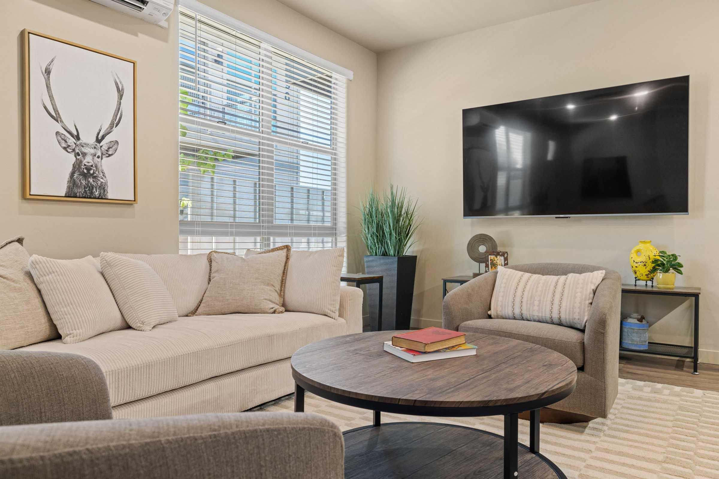 A cozy living room featuring a light-colored sofa with decorative cushions, a round coffee table stacked with books, and a flat-screen TV mounted on the wall. There's a plant in the corner and a framed artwork of a deer head on the wall, along with natural light streaming through large windows.