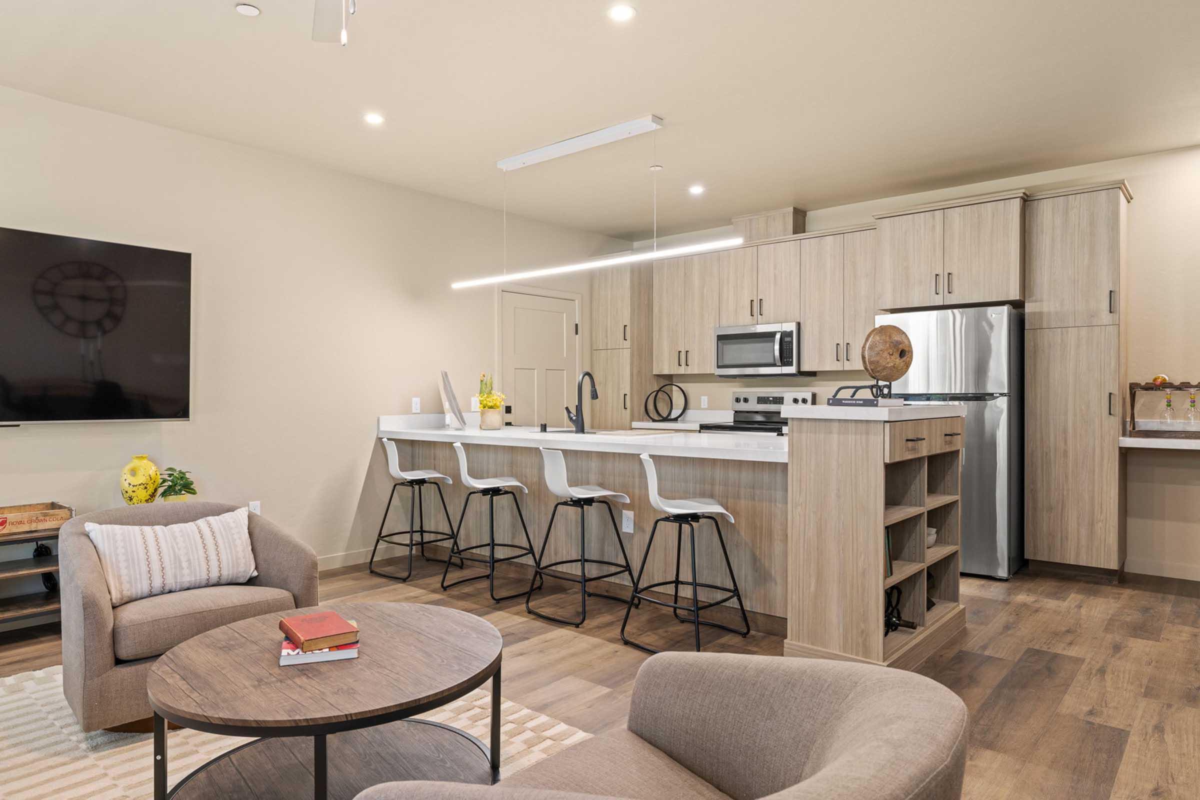 Contemporary living space featuring a modern kitchen with a bar counter and four high stools. A circular wooden coffee table sits in front of a cozy beige sofa. The kitchen has stainless steel appliances and wooden cabinetry, while a large flat-screen TV is mounted on the wall, complemented by warm lighting.