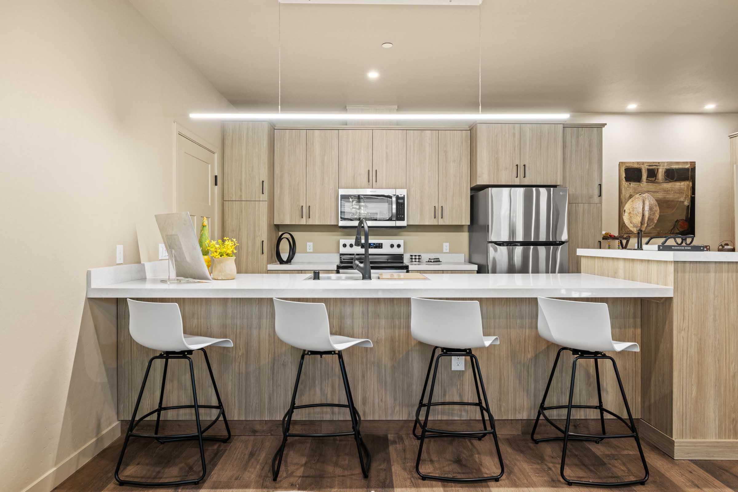 Modern kitchen with light wood cabinetry and stainless steel appliances. A spacious island features a white countertop with four sleek white bar stools. Decorative items and a small vase with yellow flowers are placed on the counter, creating a bright and inviting atmosphere.