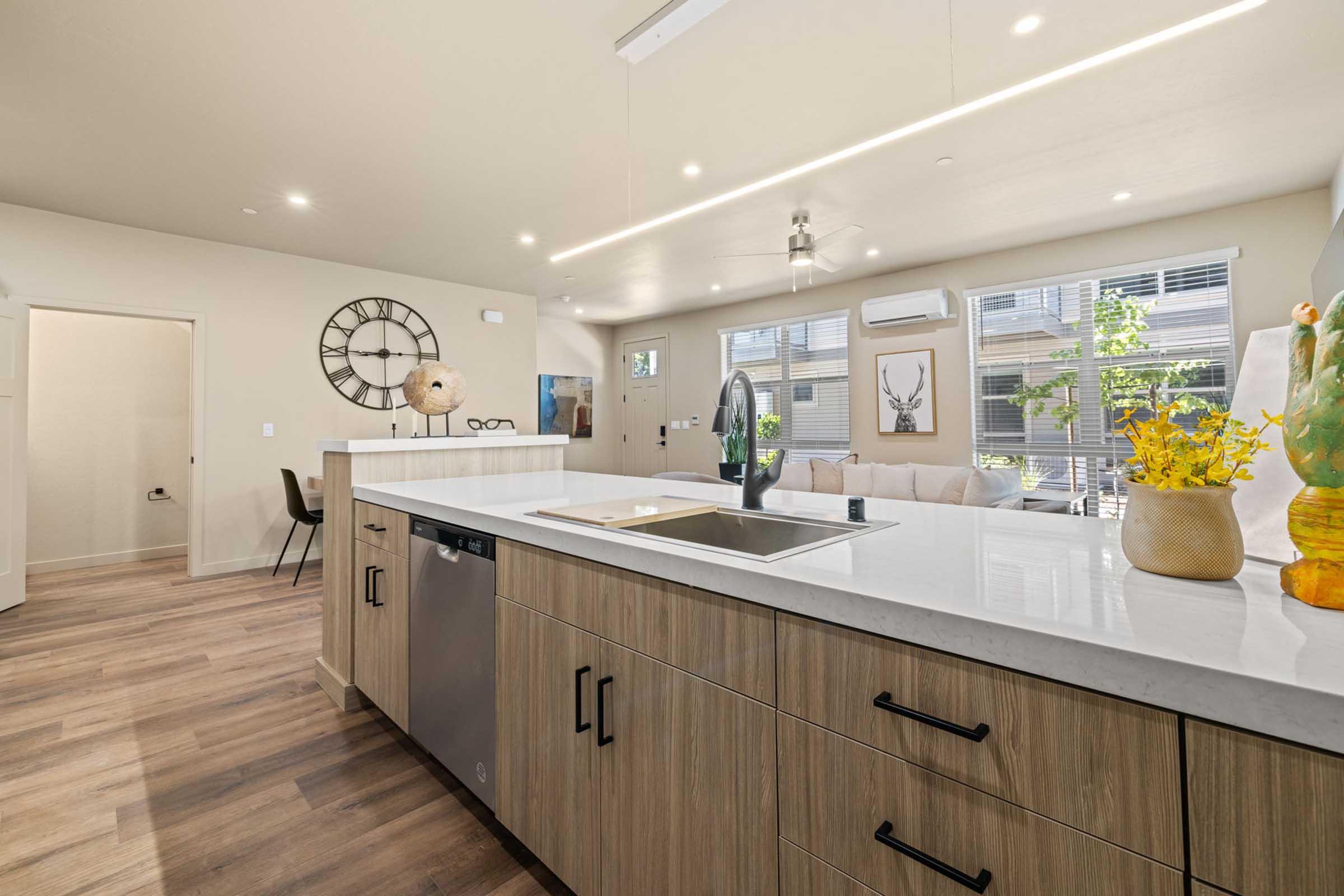 Modern kitchen featuring a central island with a sink, dishwasher, and stylish wooden cabinetry. The open layout connects to a spacious living area with large windows, a comfortable couch, and decorative elements like a globe and a vase with flowers. Brightly lit with contemporary finishing touches.