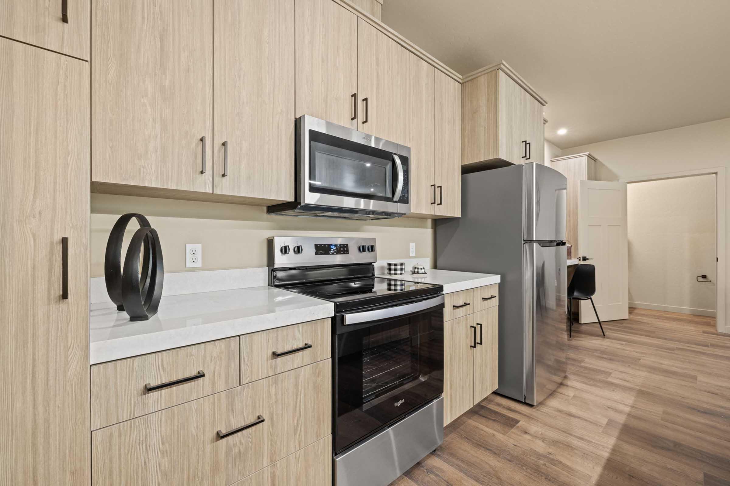 Modern kitchen featuring light wood cabinetry, stainless steel appliances including a microwave and refrigerator, a sleek oven, and a clean countertop. The space has warm wood flooring, and a cozy dining area is visible in the background.