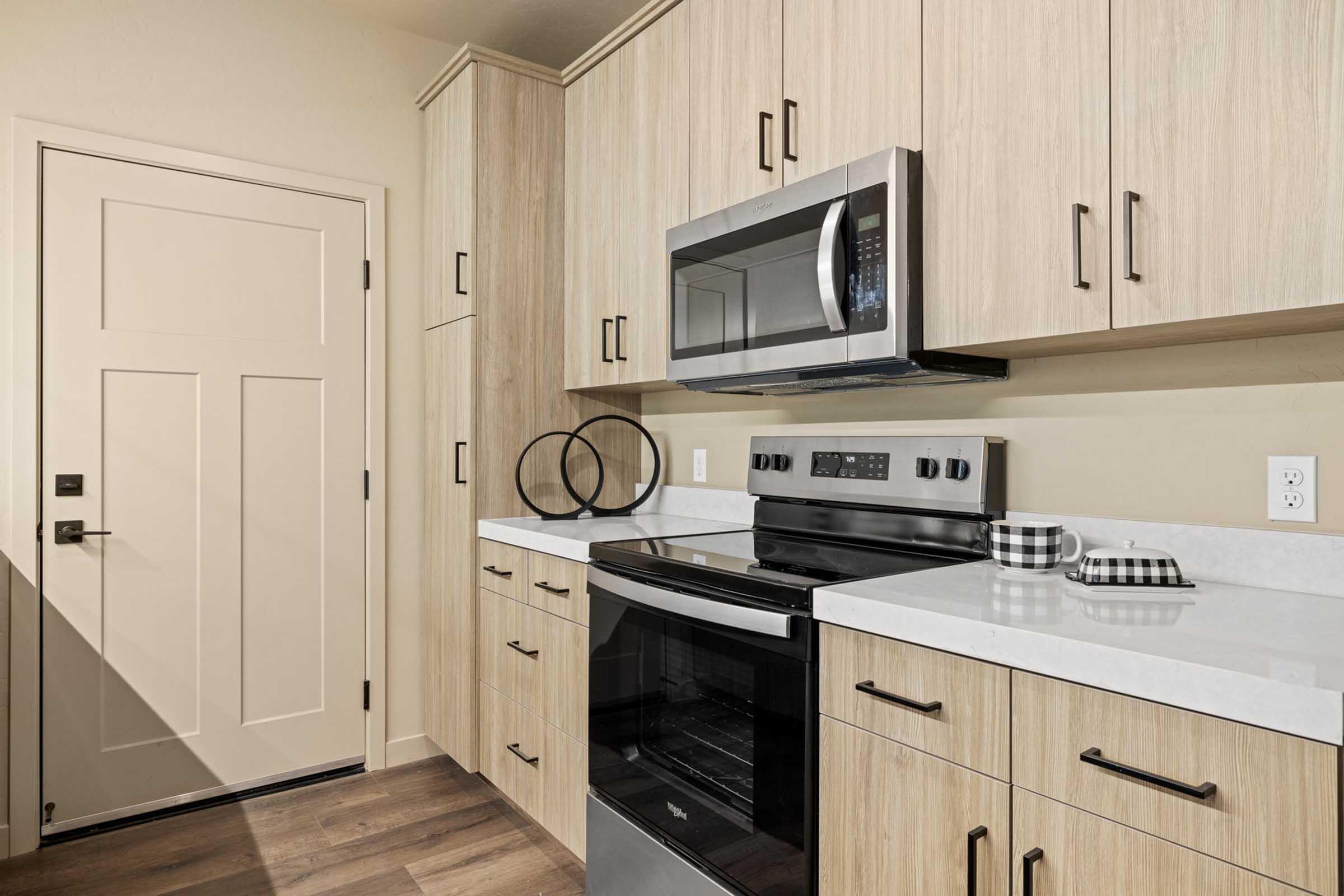 Modern kitchen featuring light wood cabinetry, a stainless steel microwave, and an electric stove. The countertops are white, with a checkered dish towel and decorative items on display. A cream-colored door is visible in the background, adding to the clean, contemporary design.