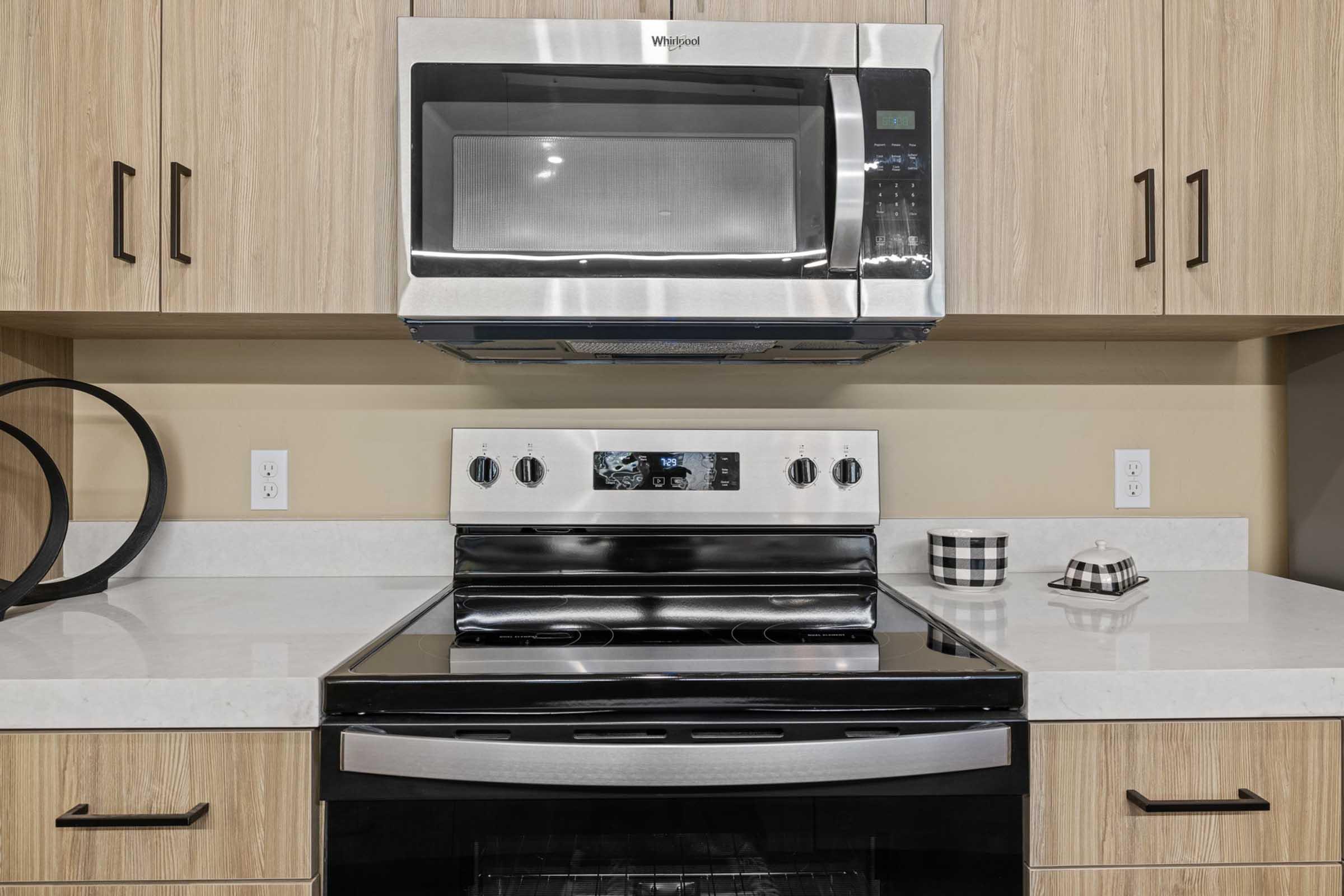 A modern kitchen featuring a stainless steel microwave mounted above a black oven with a glass top. The cabinetry is light wood, and the countertop is white. There are two wall outlets and a decorative checkered pattern canister on the counter, along with a small dish.