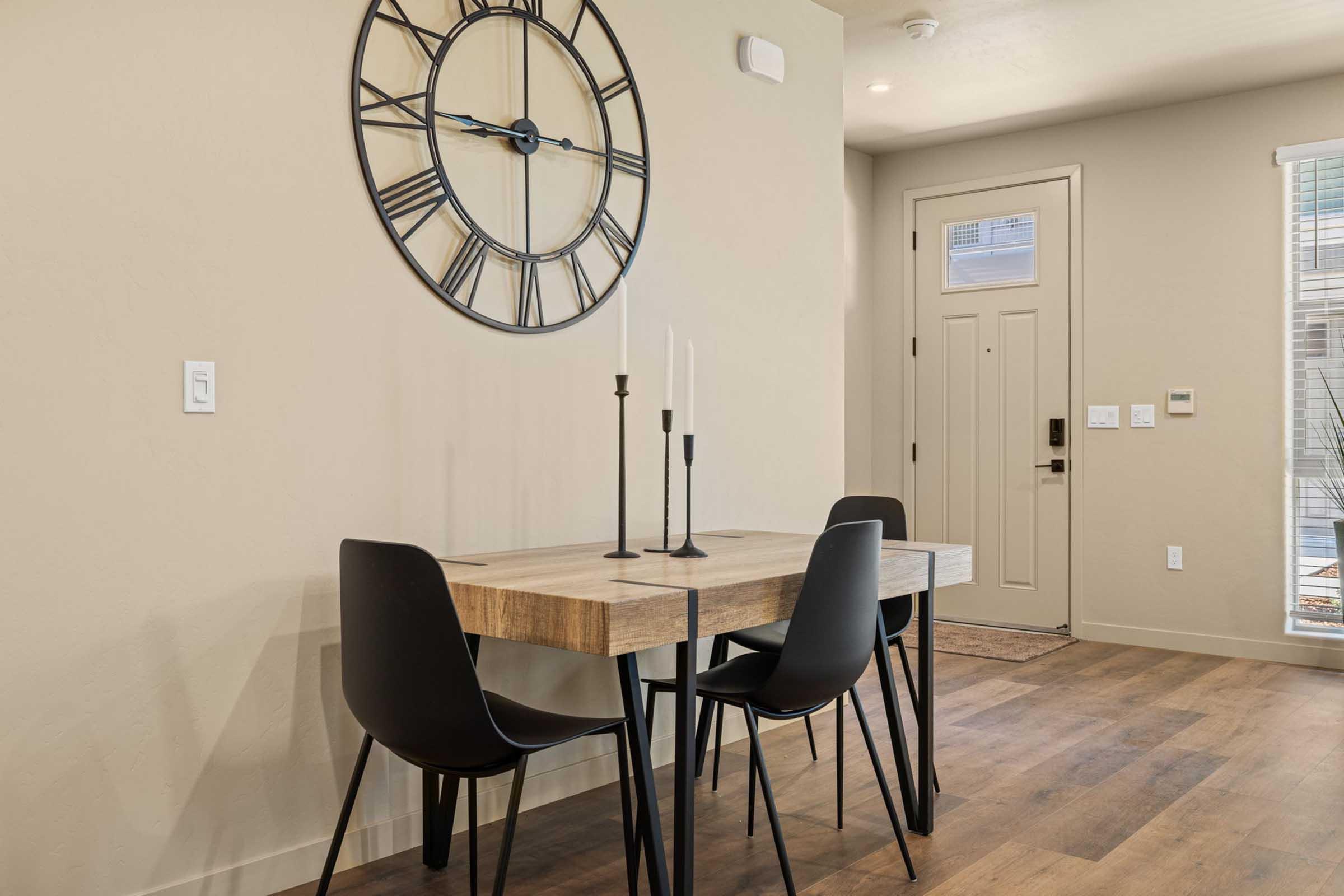 A modern dining area featuring a wooden table with black chairs, a simple candle holder, and a large wall clock. The room has neutral-colored walls and wooden flooring, with a door leading outside visible in the background. Natural light streams in through the windows.