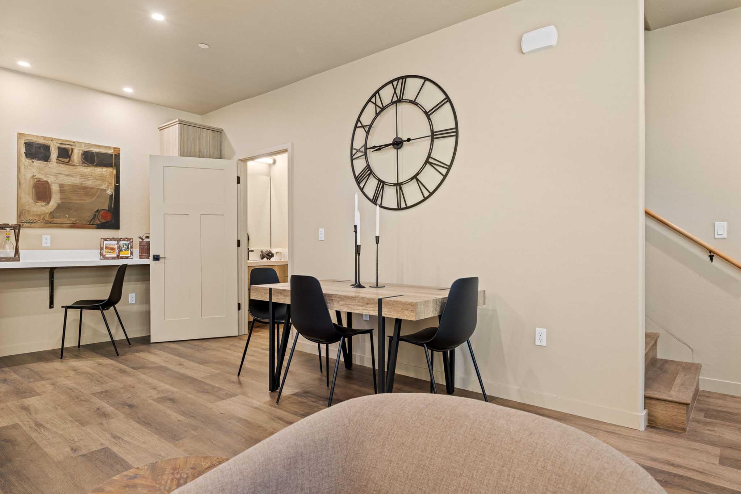 A modern interior space featuring a dining area with a round table and four black chairs. A large wall clock hangs above the table, and there’s a small workspace with a counter and stool in the background. The walls are painted in soft tones, with wooden flooring and some decorative art on display.
