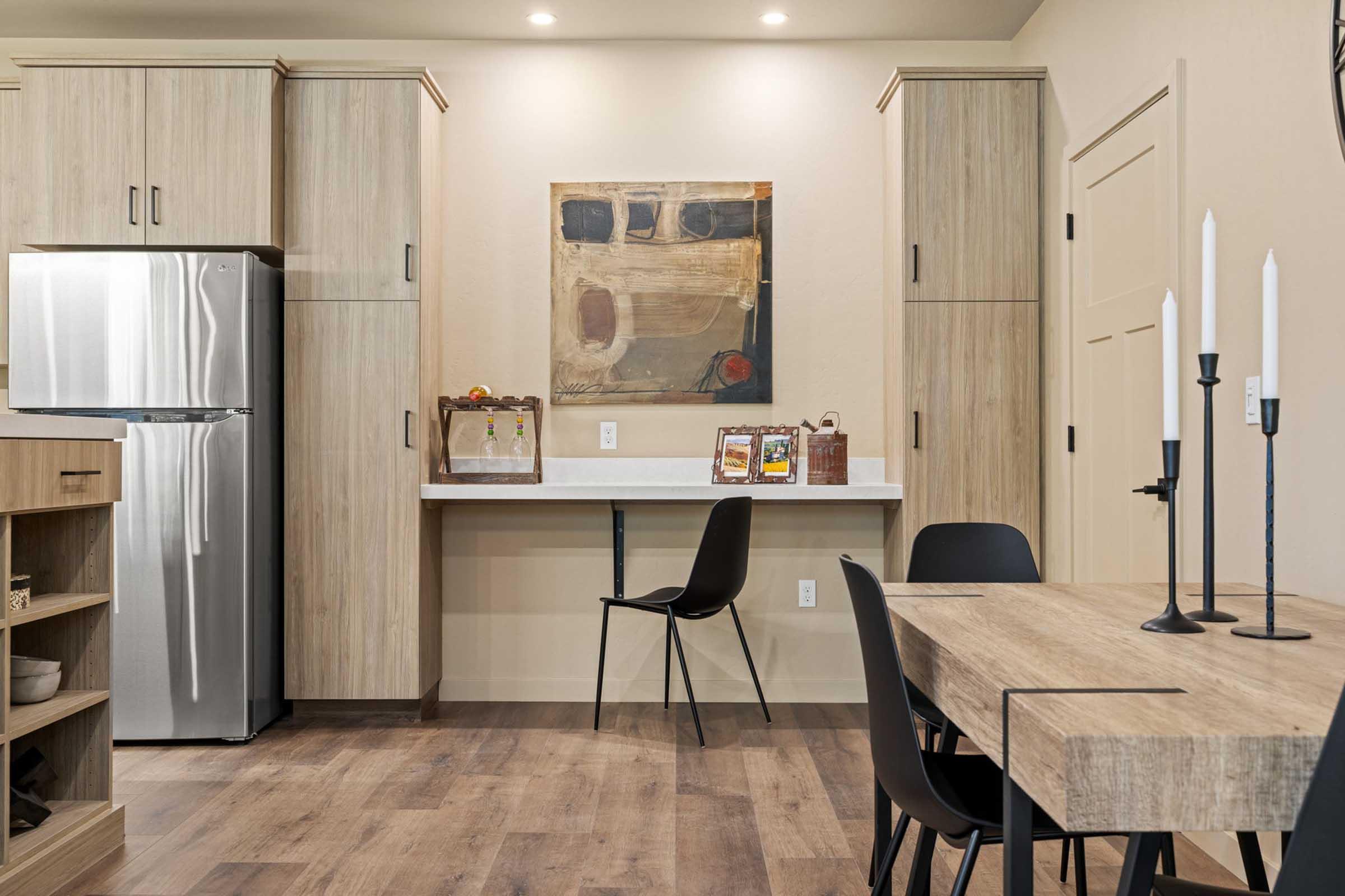 Modern kitchen with wooden cabinets, stainless steel refrigerator, and a dining table. A chair is positioned at a small desk area with artwork on the wall above. Two candlesticks are on the table, enhancing the contemporary and warm ambiance of the space.