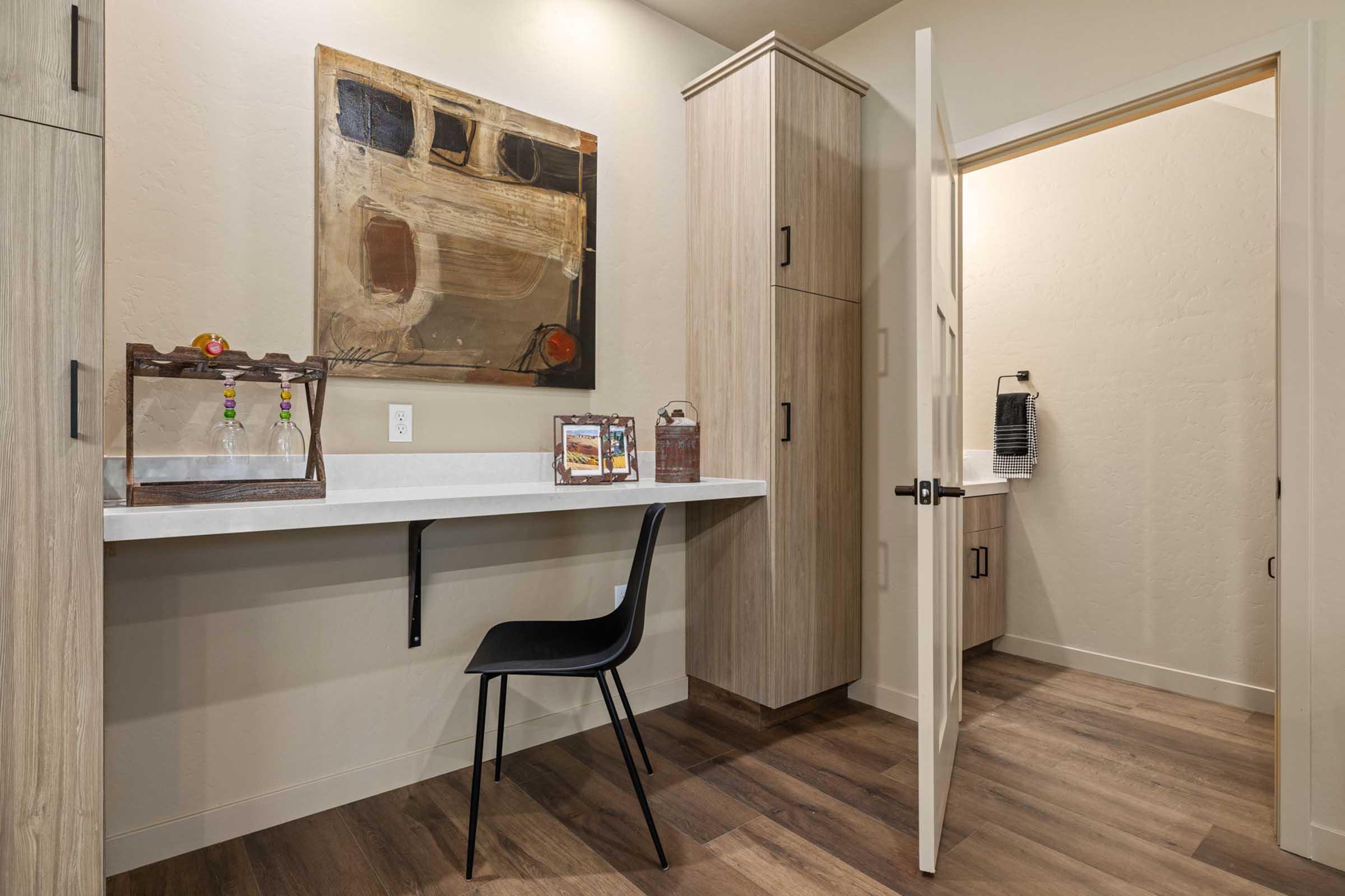 A modern workspace featuring a wall-mounted desk with a black chair, light-colored cabinetry, and a decorative painting on the wall. A doorway leads to a small room with additional storage, and the space is highlighted by warm wood flooring and soft lighting.