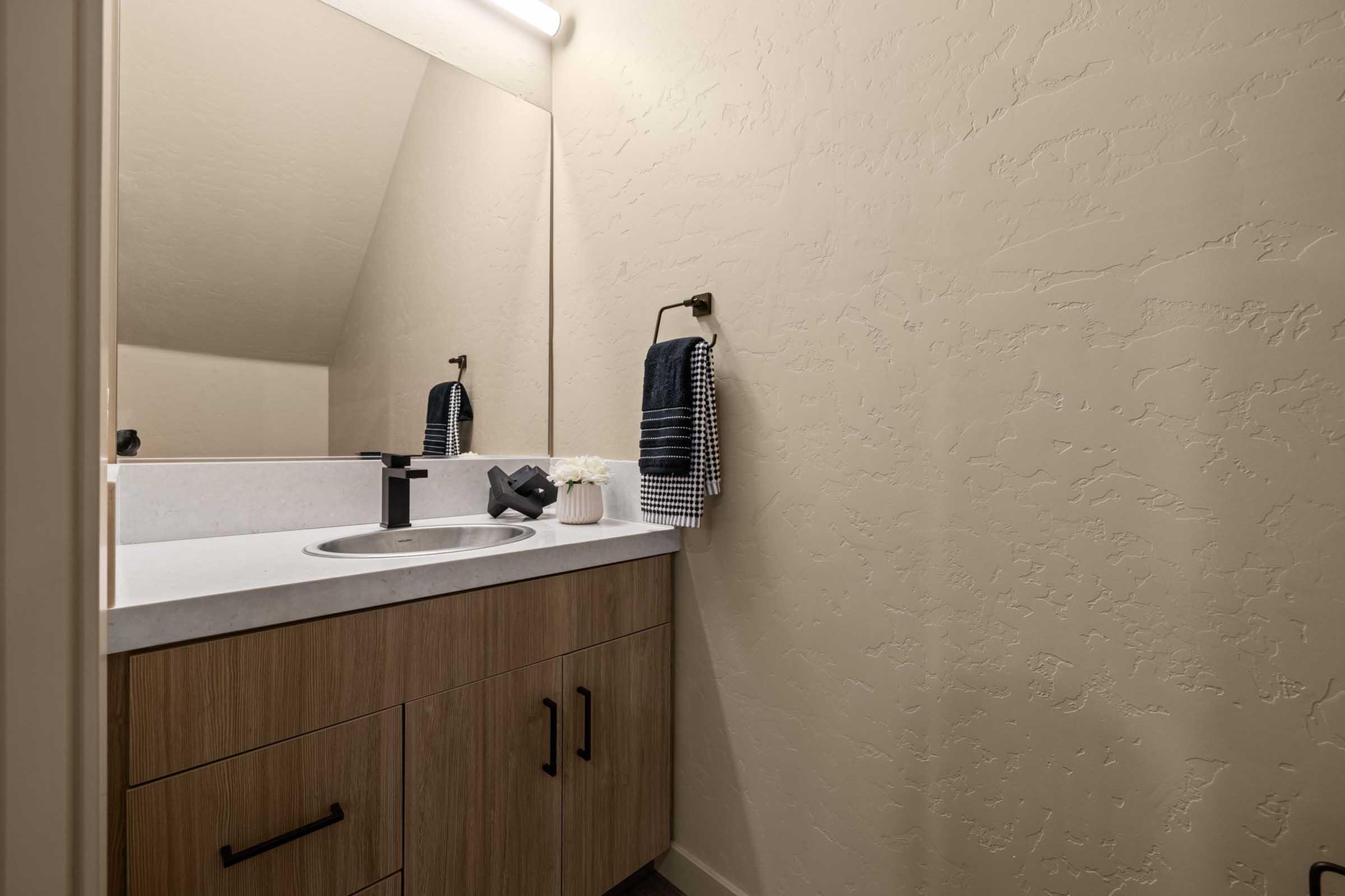 A modern bathroom featuring a countertop sink, a large mirror, and a wooden vanity with sleek black handles. A hand towel hangs on a holder next to the sink, and the walls are painted a soft beige. A light fixture provides bright lighting above the mirror, enhancing the clean, minimalist design.