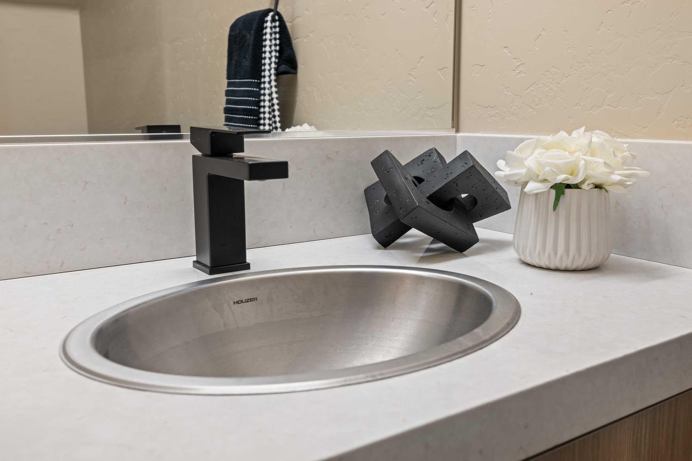 A modern bathroom sink with a sleek black faucet and a round stainless steel basin. Nearby, there is a decorative black geometric sculpture and a white ceramic vase filled with artificial flowers. A black and white striped hand towel hangs on the wall in the background.