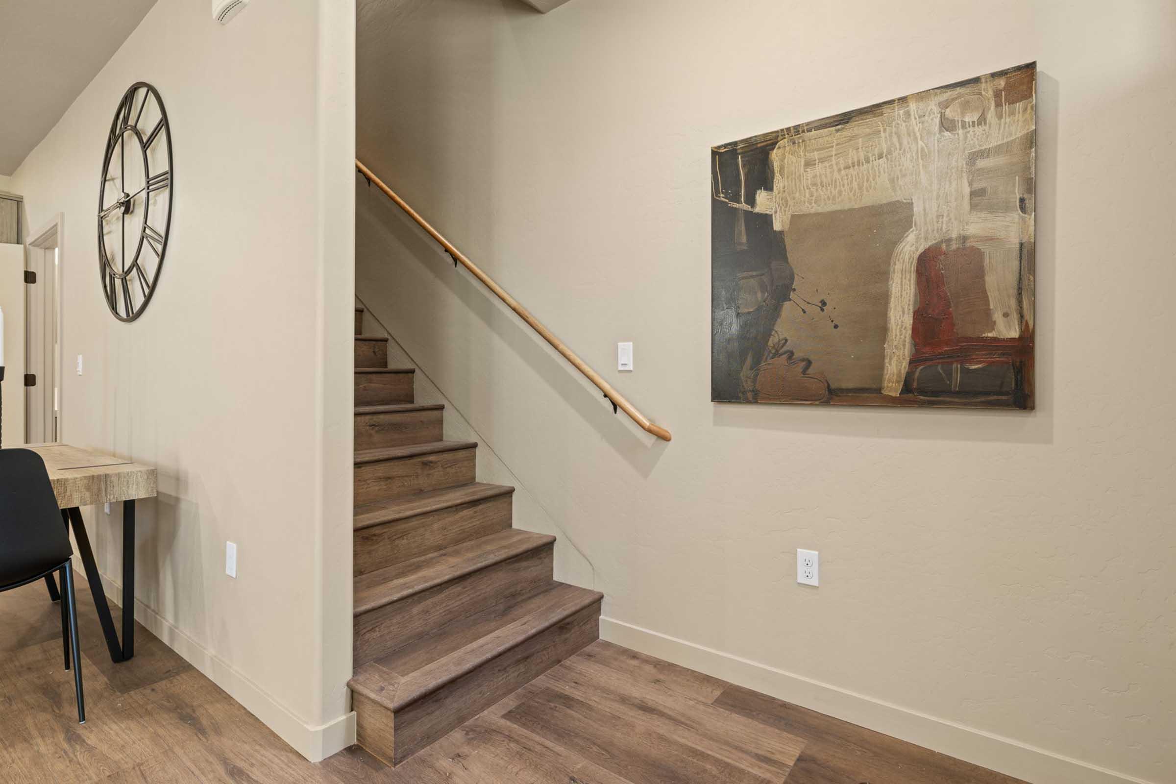 A modern hallway featuring a set of wooden stairs leading to an upper level. On the wall, there is a large abstract painting. A round wall clock is also visible. The floor has a warm wooden finish, and there is a small table with a black chair nearby. The overall color scheme is neutral and contemporary.