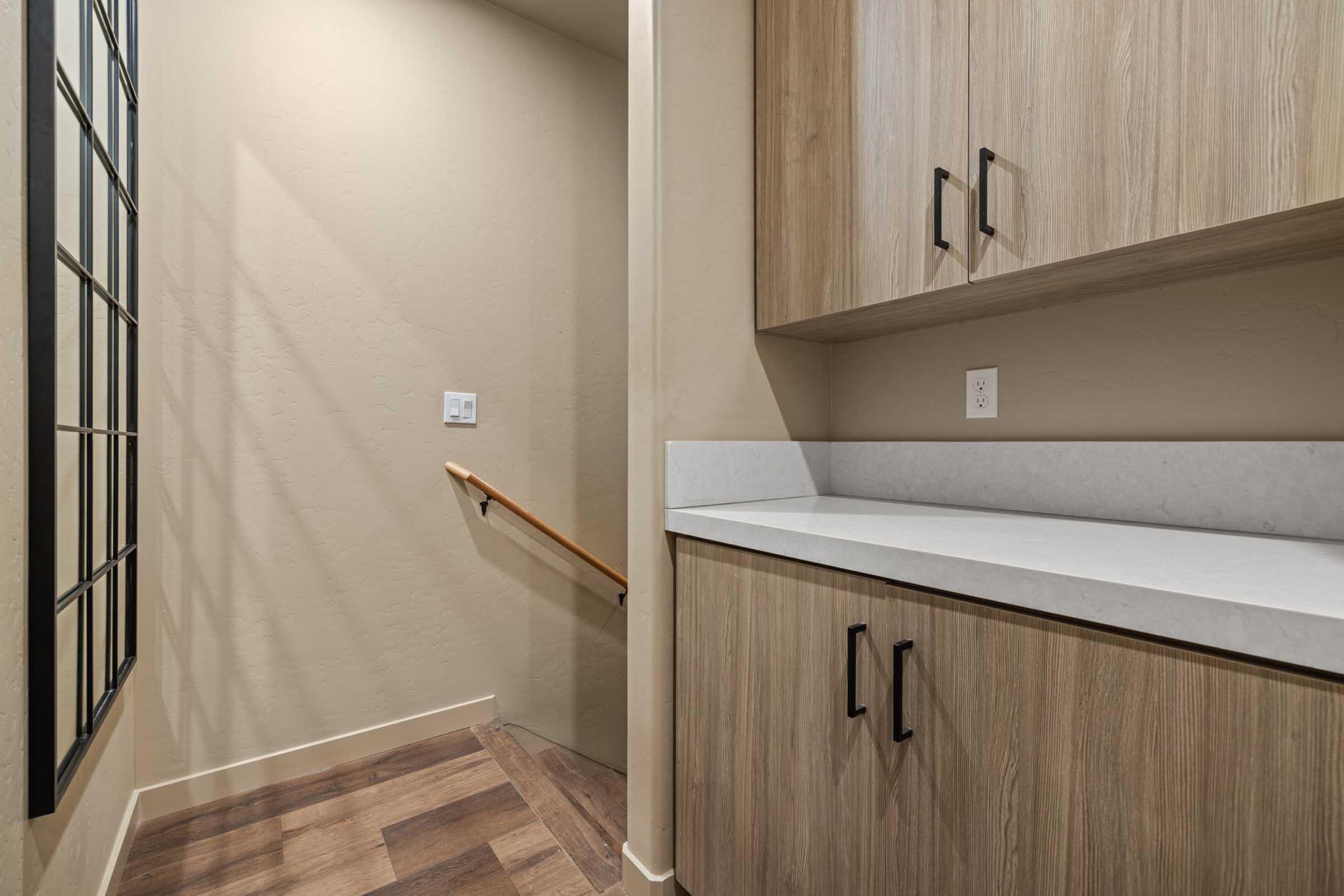 A partially visible stairway leading to an upper level, with a light-colored wall and wooden handrail. To the right, there's a countertop with a white surface and wooden cabinets below. The floor features dark wood planks, and a window with a grid pattern is seen on the left.