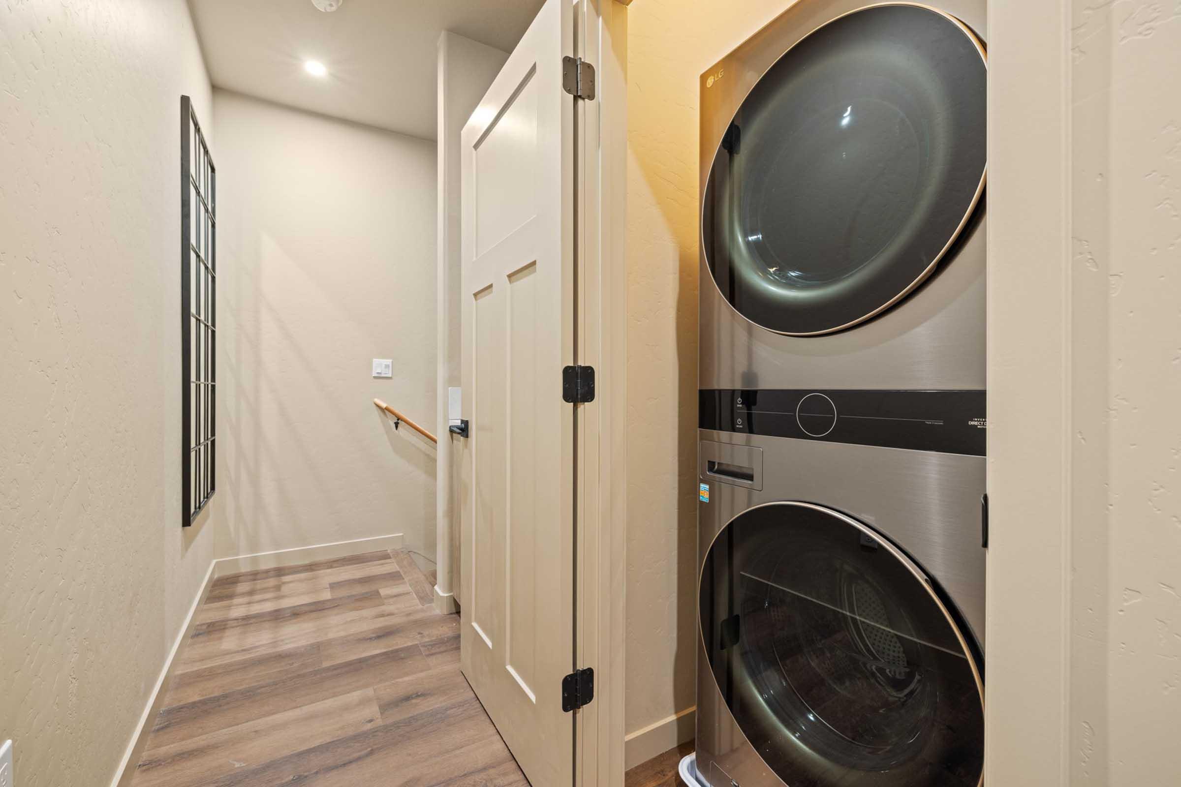 A narrow hallway leading to a door, with a stacked washer and dryer unit positioned against the wall. The floor features light wood-like flooring, and the walls are painted a soft neutral color. A set of stairs is visible at the end of the hallway, along with a window.