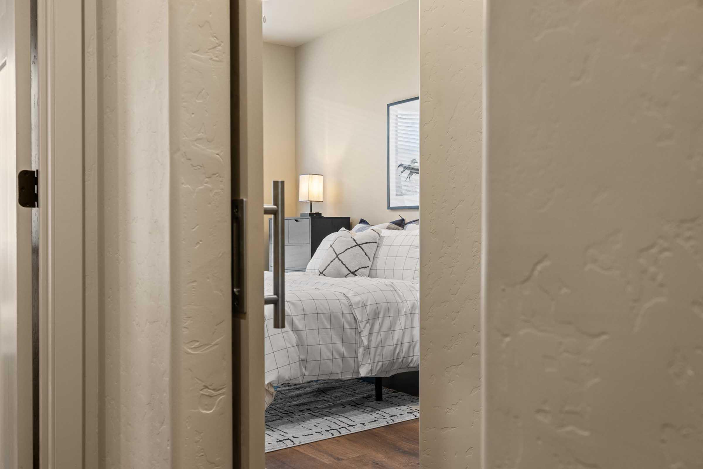 A cozy bedroom view through a slightly open door. The room features a neatly made bed with a white and gray plaid duvet, accented pillows, and a bedside lamp. A decorative rug is visible on the wooden floor, and a framed picture hangs on the wall, adding to the inviting atmosphere.