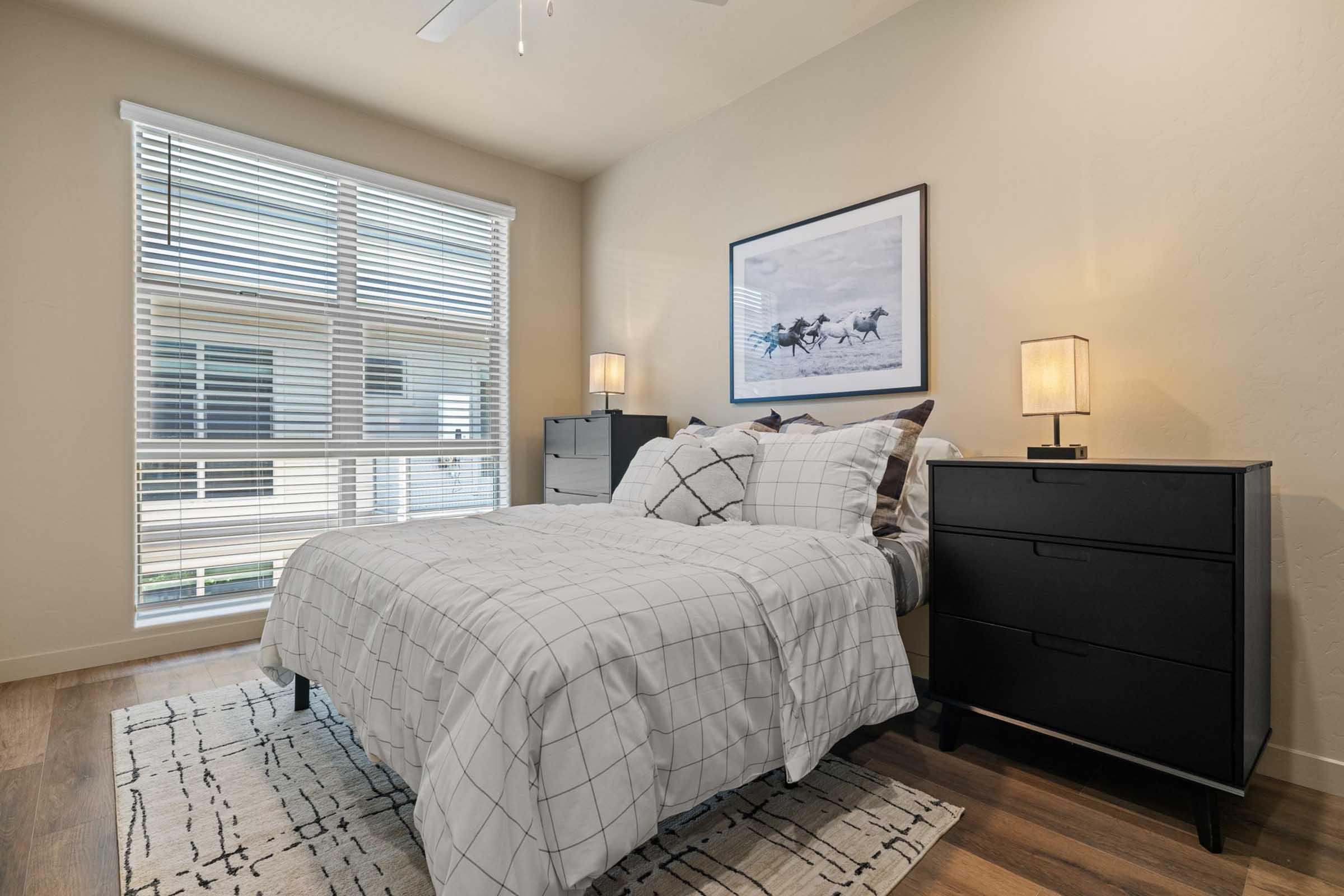 A cozy bedroom featuring a queen-sized bed with a patterned gray and white comforter, two bedside lamps, and a modern black dresser. Large windows let in natural light, complemented by sheer blinds. A minimalistic rug adds warmth to the wooden floor, creating a snug and inviting atmosphere.
