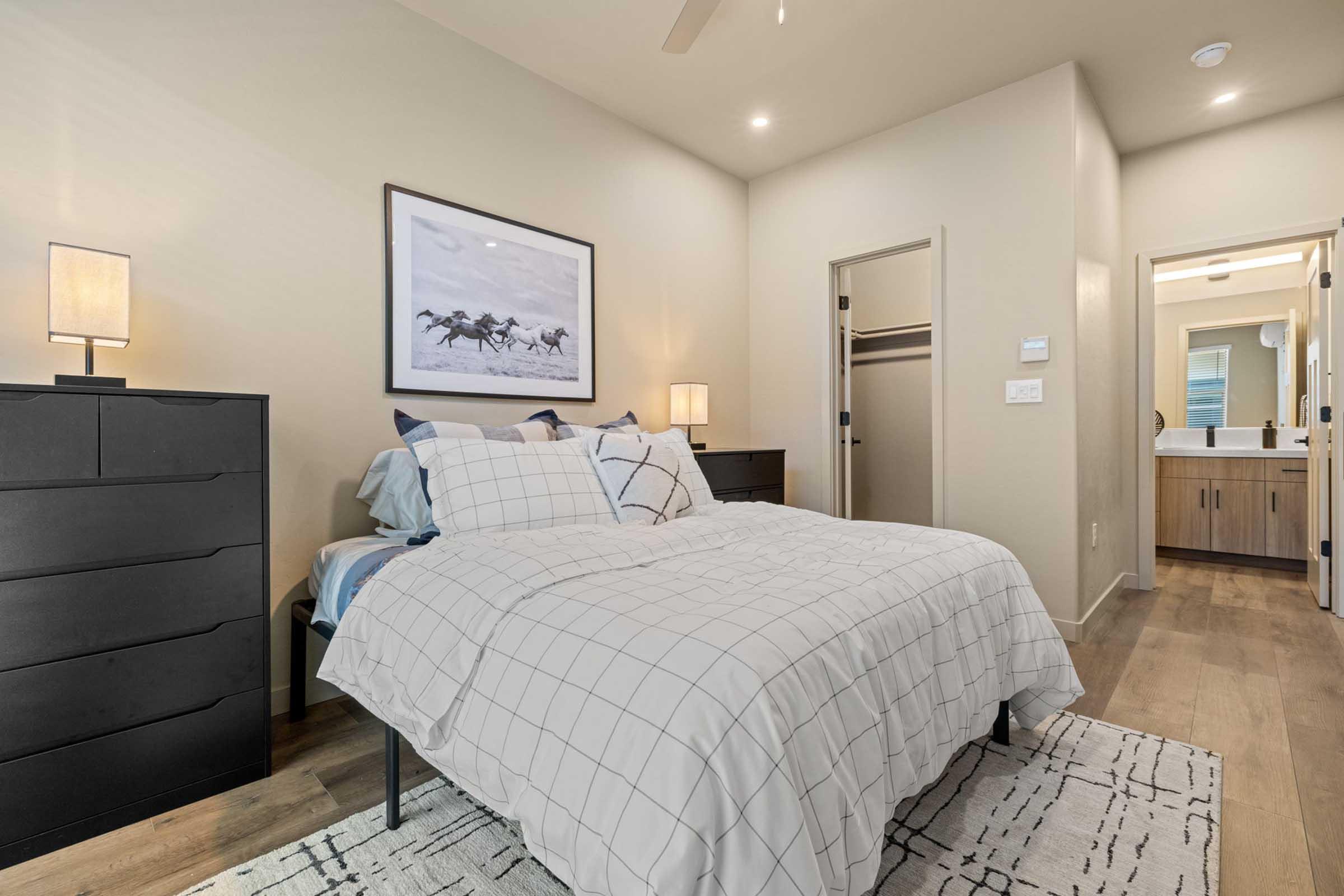 A modern bedroom featuring a bed with a white plaid duvet, two bedside lamps, and a large framed artwork on the wall. There's a black dresser, and a closet door is slightly ajar. In the background, a doorway leads to a bathroom with wooden cabinetry. The room has neutral-colored walls and wooden flooring.