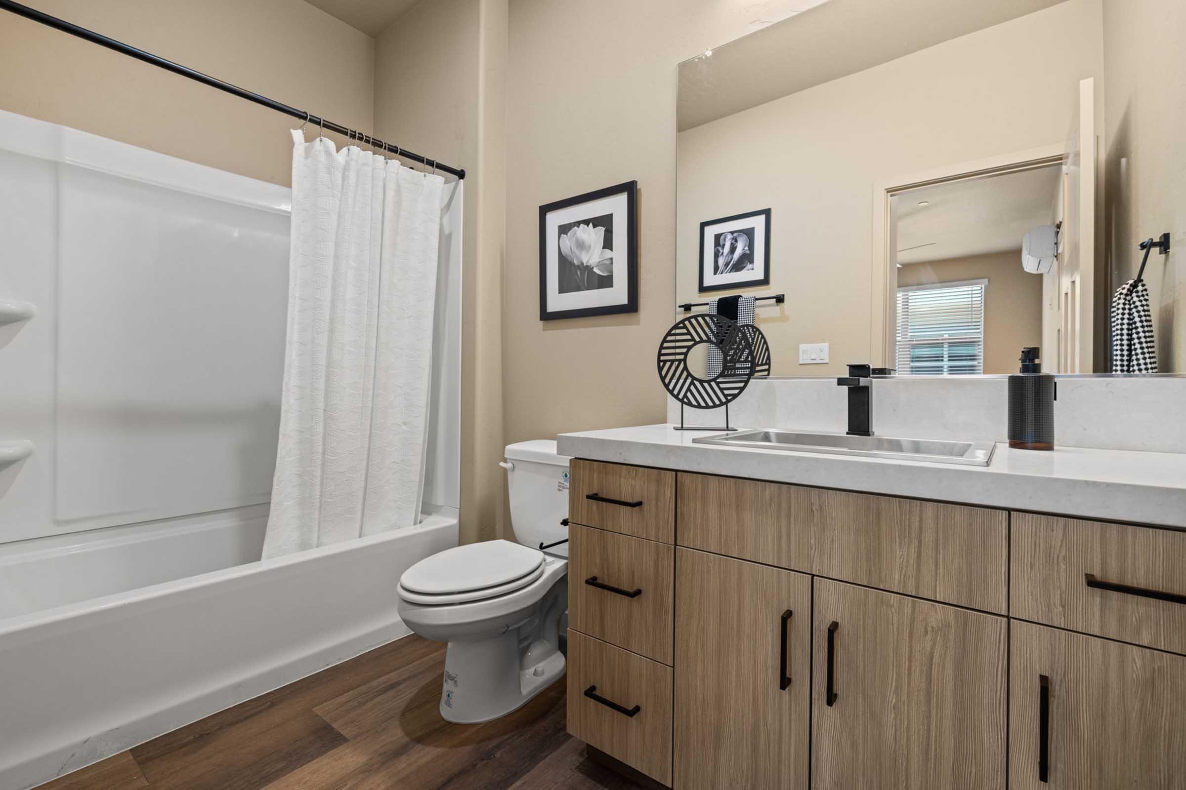 A modern bathroom featuring a shower/tub combination with a white curtain, a rectangular sink with sleek black faucets, wooden cabinetry, and a black fan. The walls are painted in a neutral tone, and there are two framed pictures on the wall, adding a decorative touch to the space.