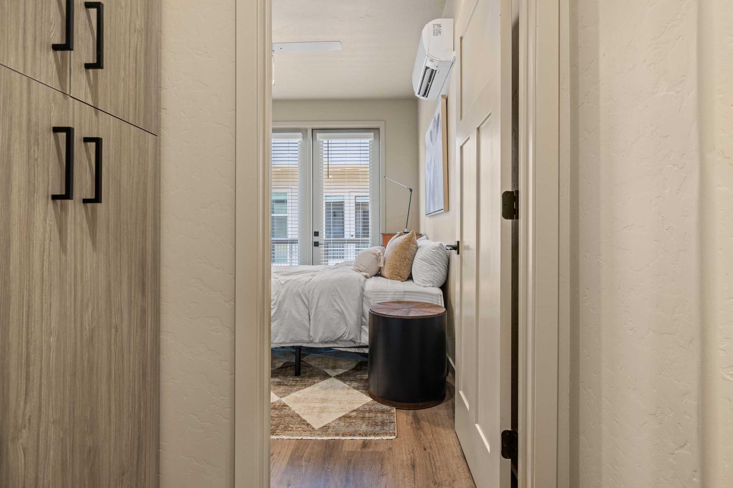 A view through a doorway into a cozy bedroom featuring a bed with white linens, a bedside table, and a decorative rug. The room has large windows allowing natural light and is decorated in neutral tones, creating a warm and inviting atmosphere.