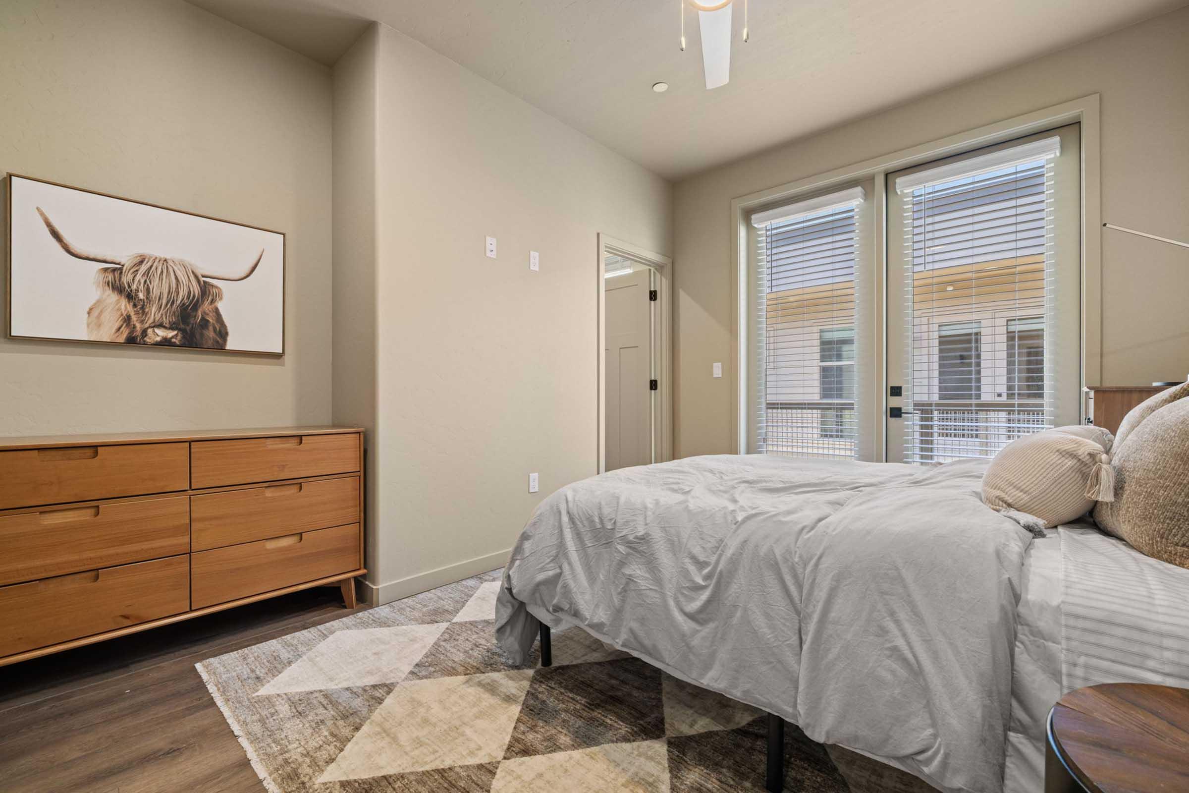 A modern bedroom featuring a neatly made bed with a light gray comforter, a large dresser made of wood, and a decorative wall art of a longhorn cow. Natural light streams in through large windows with blinds. The floor has a stylish area rug, and the walls are painted in a soft neutral color.
