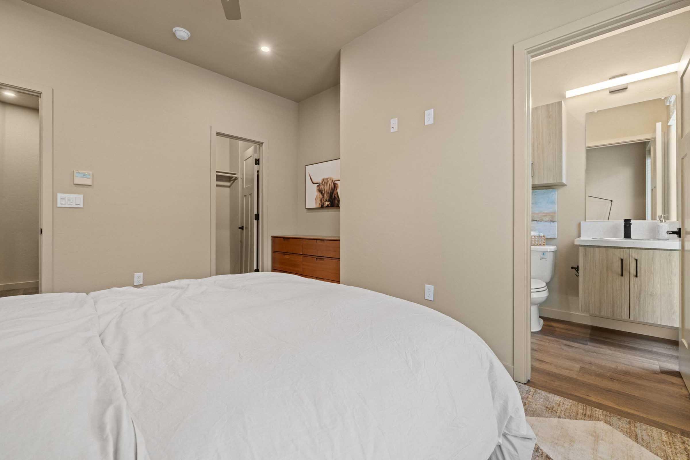 A spacious bedroom with a neatly made bed in the foreground. To the right, there is an open bathroom door revealing a modern bathroom setup. A closet door is slightly ajar, and a wooden dresser is visible against the wall. The walls are painted a light color, giving the space an airy feel.