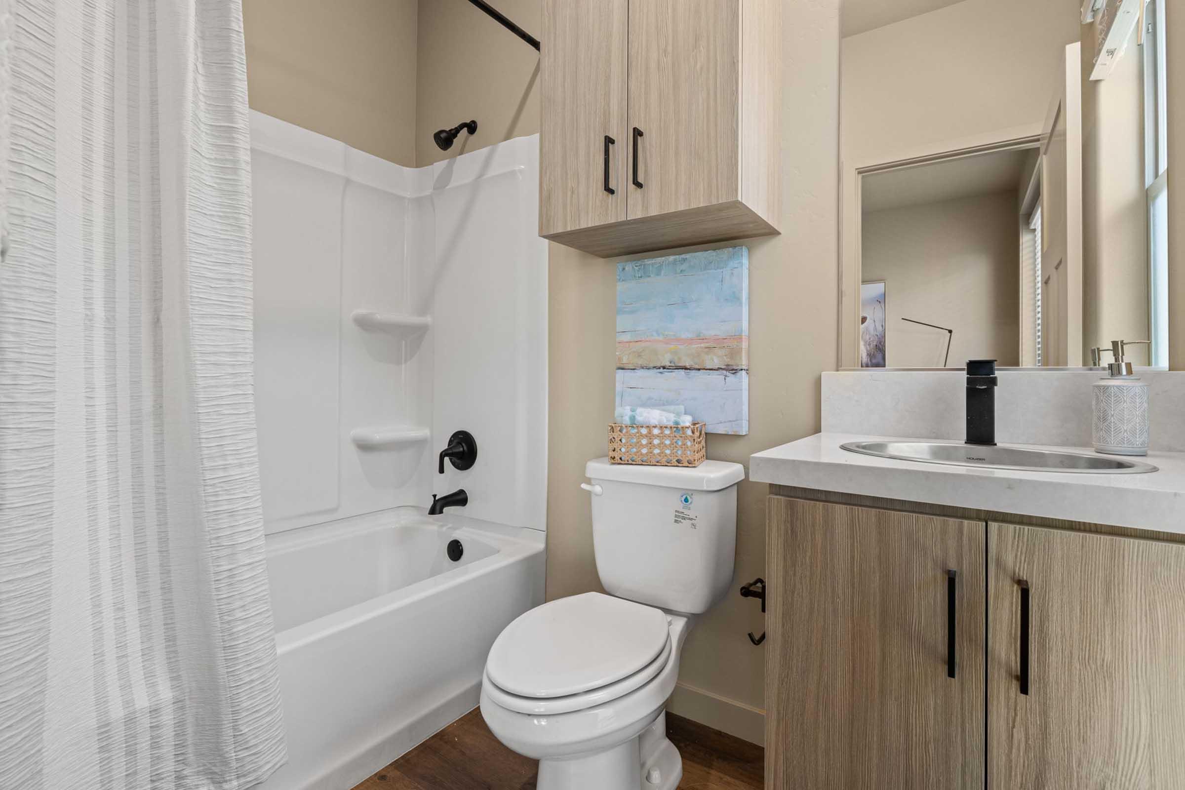 A modern bathroom featuring a white bathtub with a shower, black fixtures, and a light wood vanity. The space includes a mirror, a decorative piece on the wall, and a neatly organized bathroom area with a shower curtain and sink. Bright natural light enters through a window.