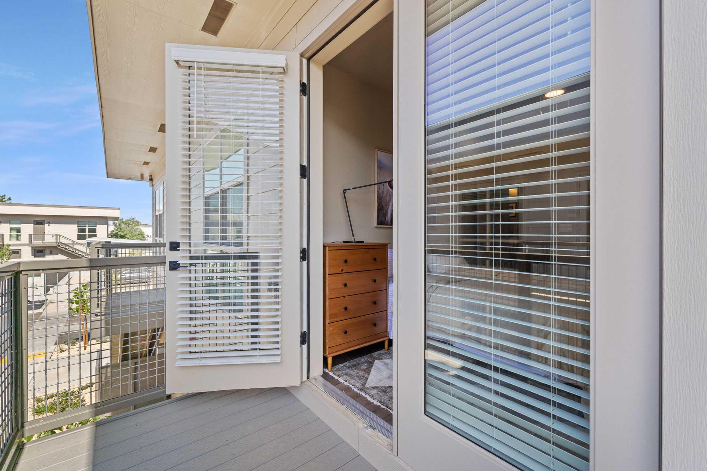 A view from a modern balcony with double doors open, showcasing a stylish interior space with light-colored walls and a wooden dresser. The balcony features a sleek railing, with a glimpse of buildings and greenery in the background on a sunny day.