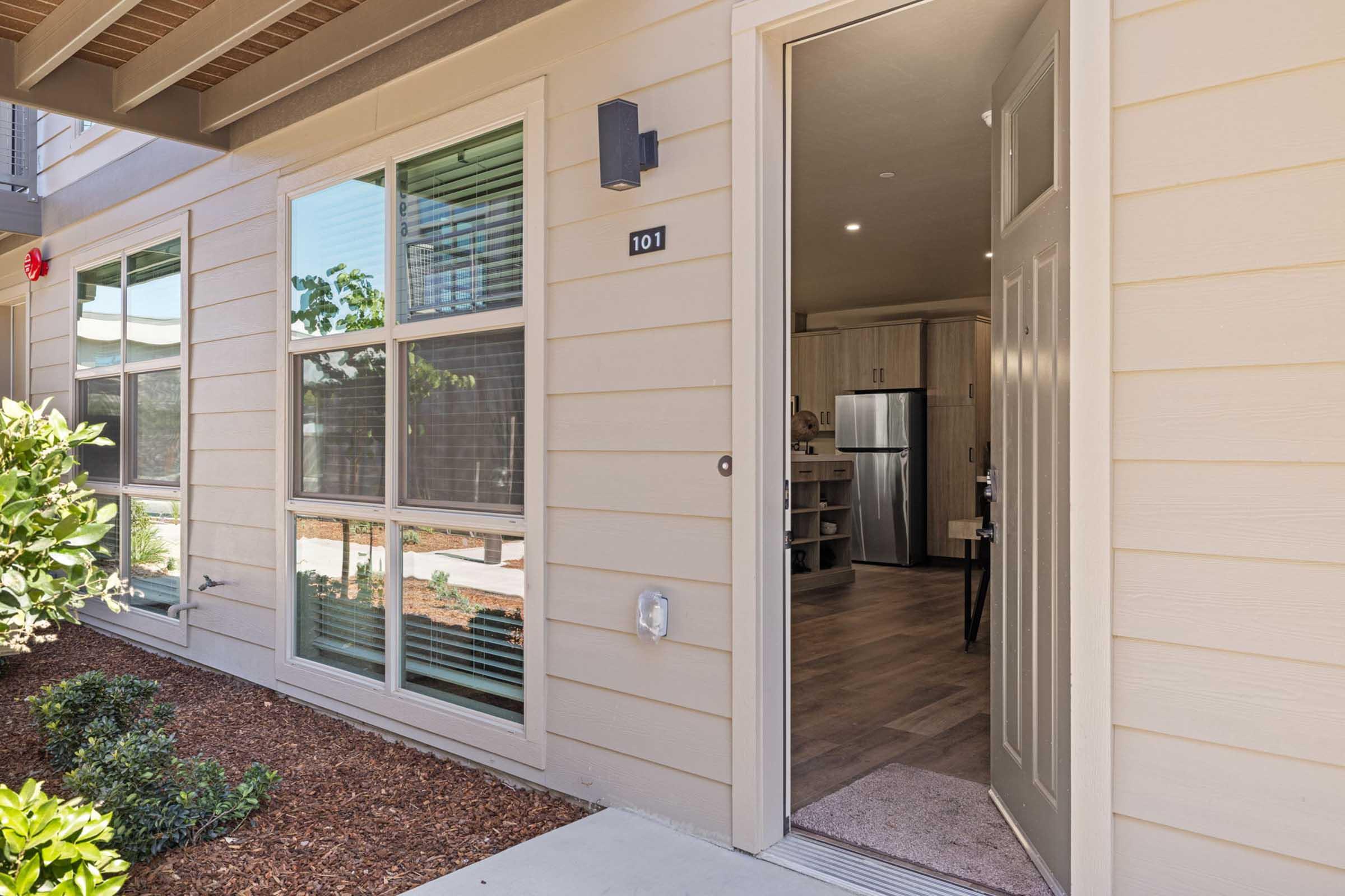 View of an open apartment door with the number 101 displayed. The entrance shows a well-lit interior with a glimpse of a kitchen and living area. Outside, there are landscaped shrubs and a pathway leading to the door. Natural light filters through the windows, creating an inviting atmosphere.
