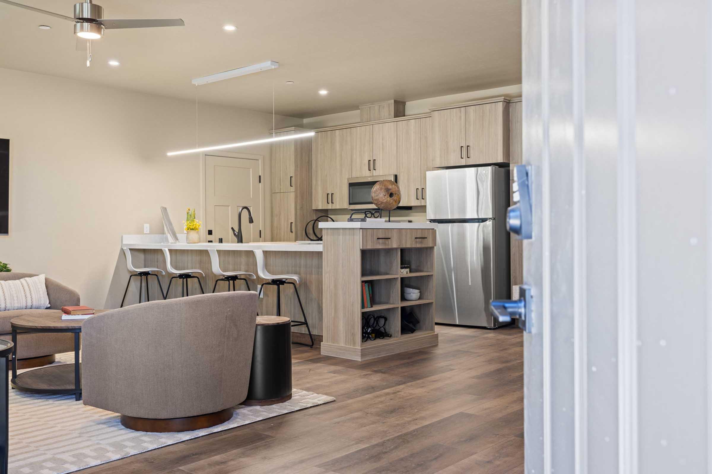Modern kitchen and living area featuring a sleek design with wood cabinetry, stainless steel appliances, and a large island with seating. The space is illuminated with ceiling lights and natural light, showcasing a comfortable seating arrangement and minimalistic decor to create an inviting atmosphere.