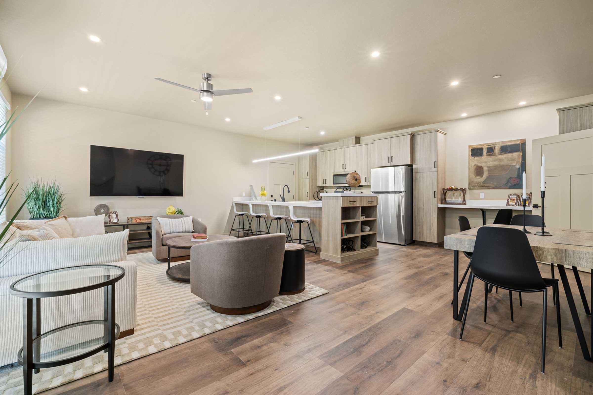 Modern open-concept living area featuring a comfortable seating arrangement with two armchairs, a sofa, and a coffee table. A large wall-mounted TV is visible, along with a dining table and stylish bar stools at a kitchen island. The space has wooden flooring and neutral-colored walls, creating a bright and inviting atmosphere.