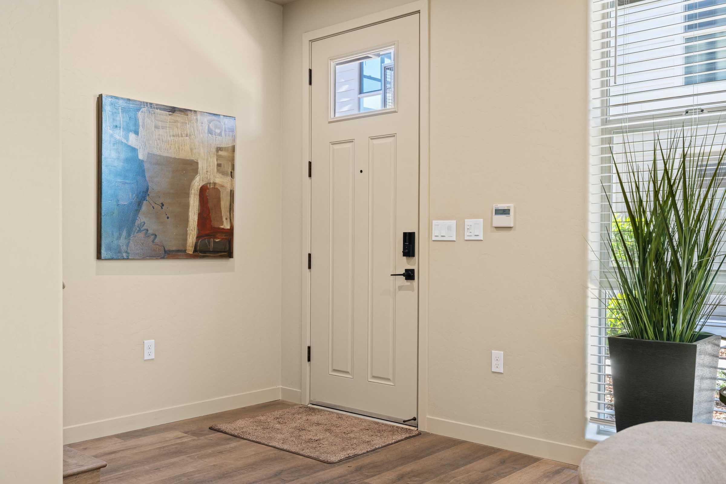 A bright entryway featuring a beige wall with a modern abstract painting, a closed door with a black handle, a welcome mat, and a large potted plant in the corner. Natural light streams in through a window beside the door, enhancing the open and inviting atmosphere.