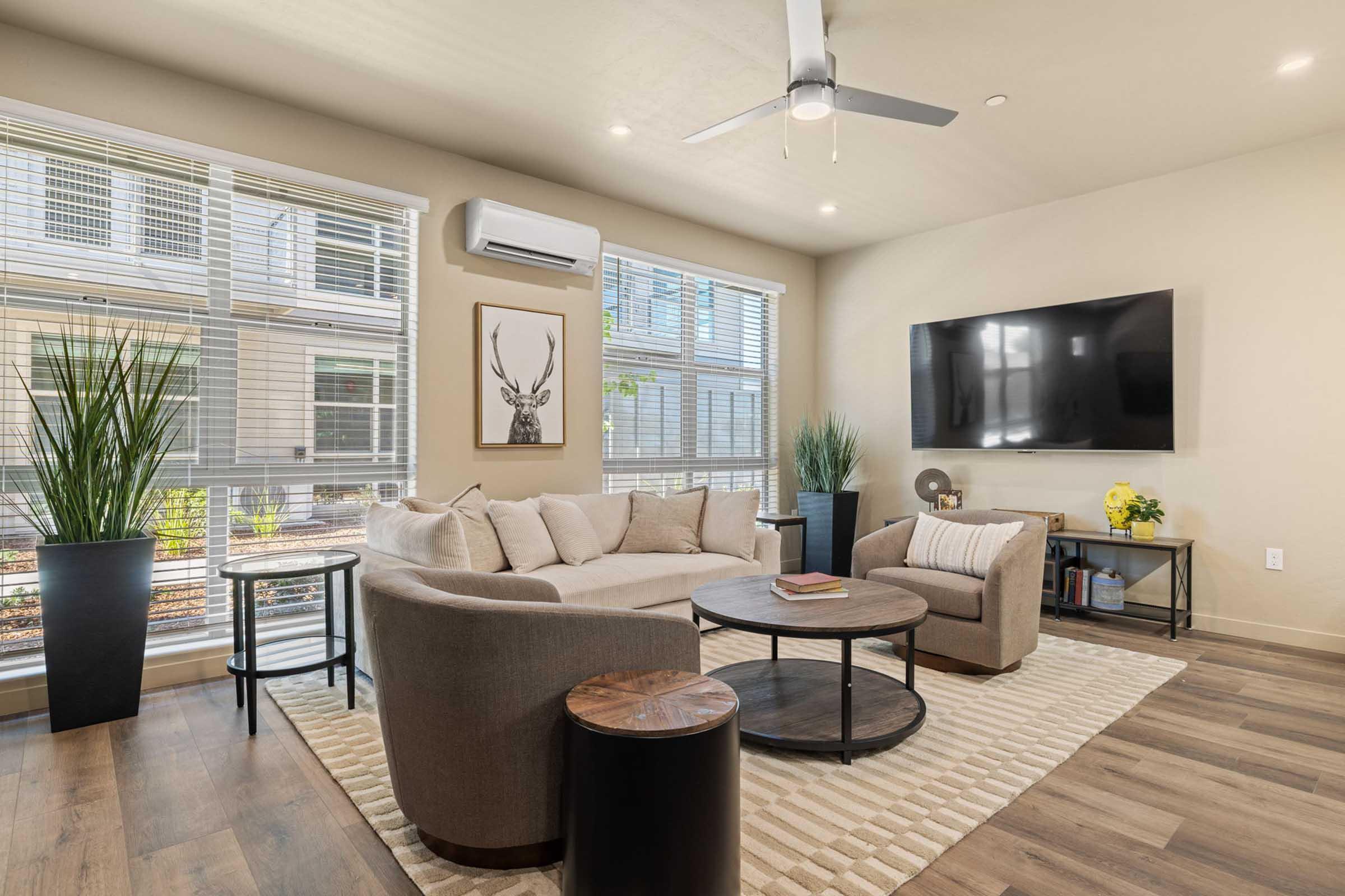 A cozy living room featuring a light-colored sectional sofa, a round coffee table, and a side table. Large windows allow natural light to fill the space, with decorative plants in the corners. A flat-screen TV is mounted on the wall, and a framed artwork is displayed. The room has a modern, inviting atmosphere.