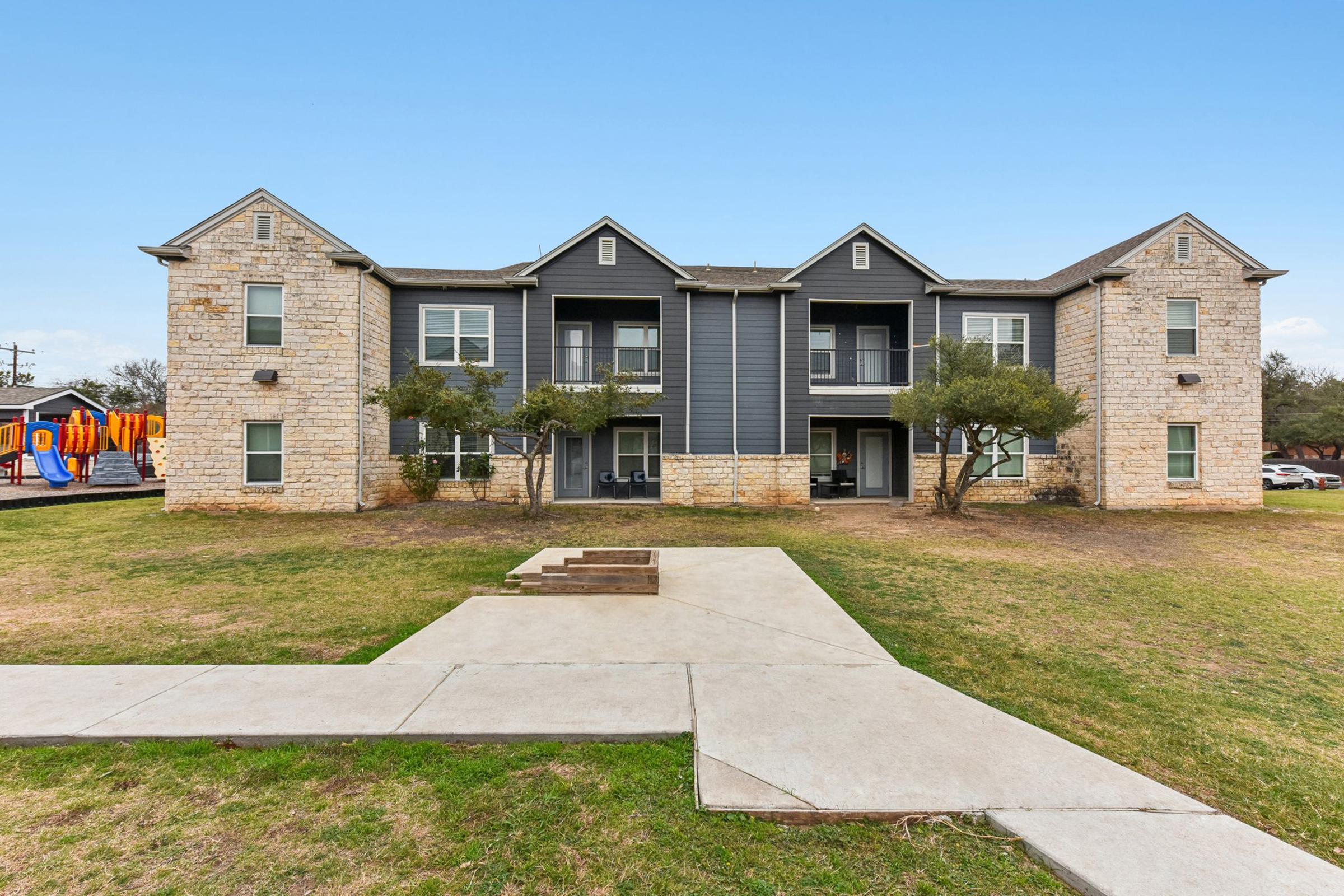 A two-story stone and gray wood apartment building with multiple balconies. The building is surrounded by a grassy area and has a walkway leading to it. In the background, there's an outdoor play area with colorful equipment. The sky is clear and blue.