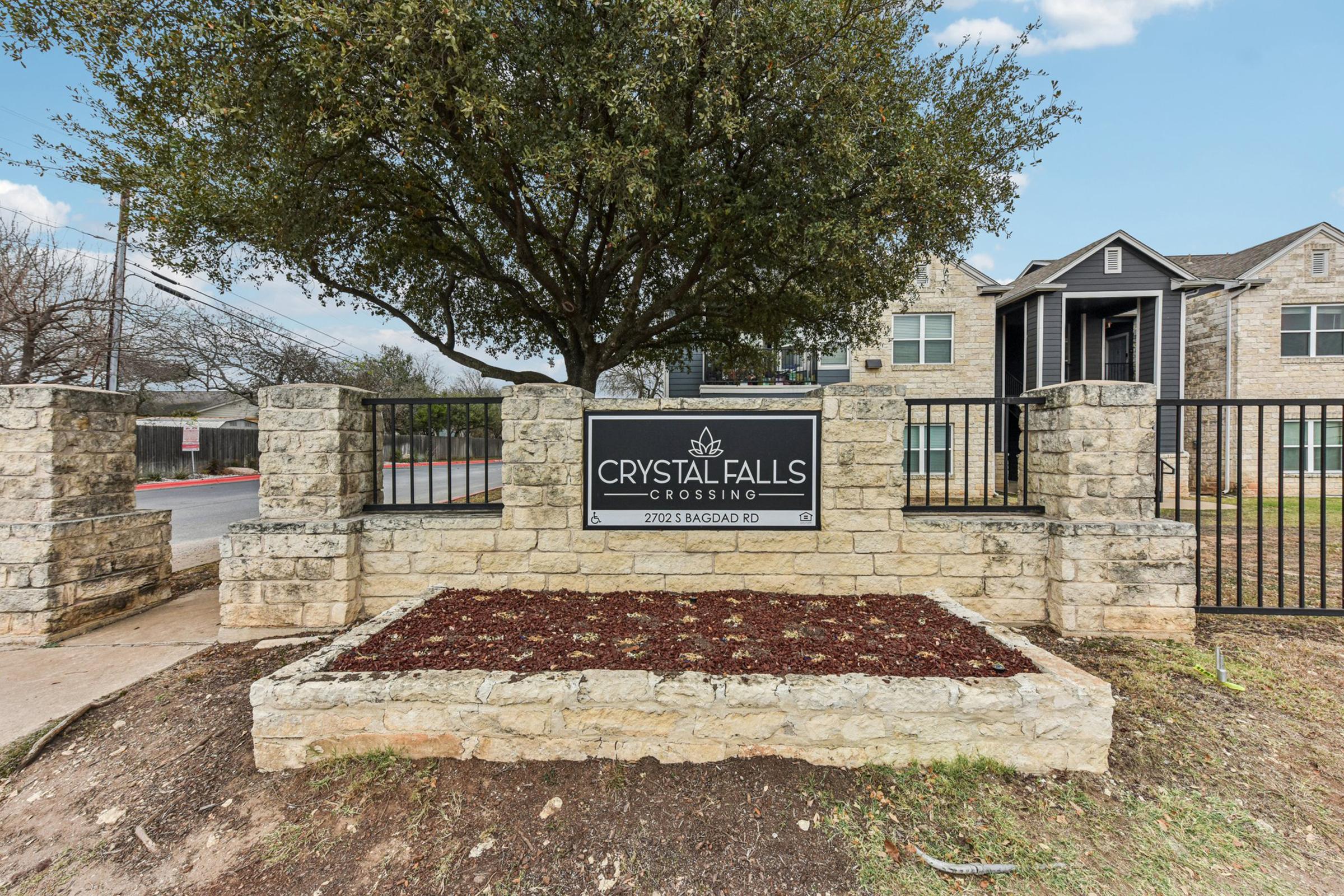 Sign for Crystal Falls Crossing, located at 2112 Bagdad Rd. The sign is surrounded by a stone wall with mulch, and there are trees and residential buildings visible in the background, under a partly cloudy sky.