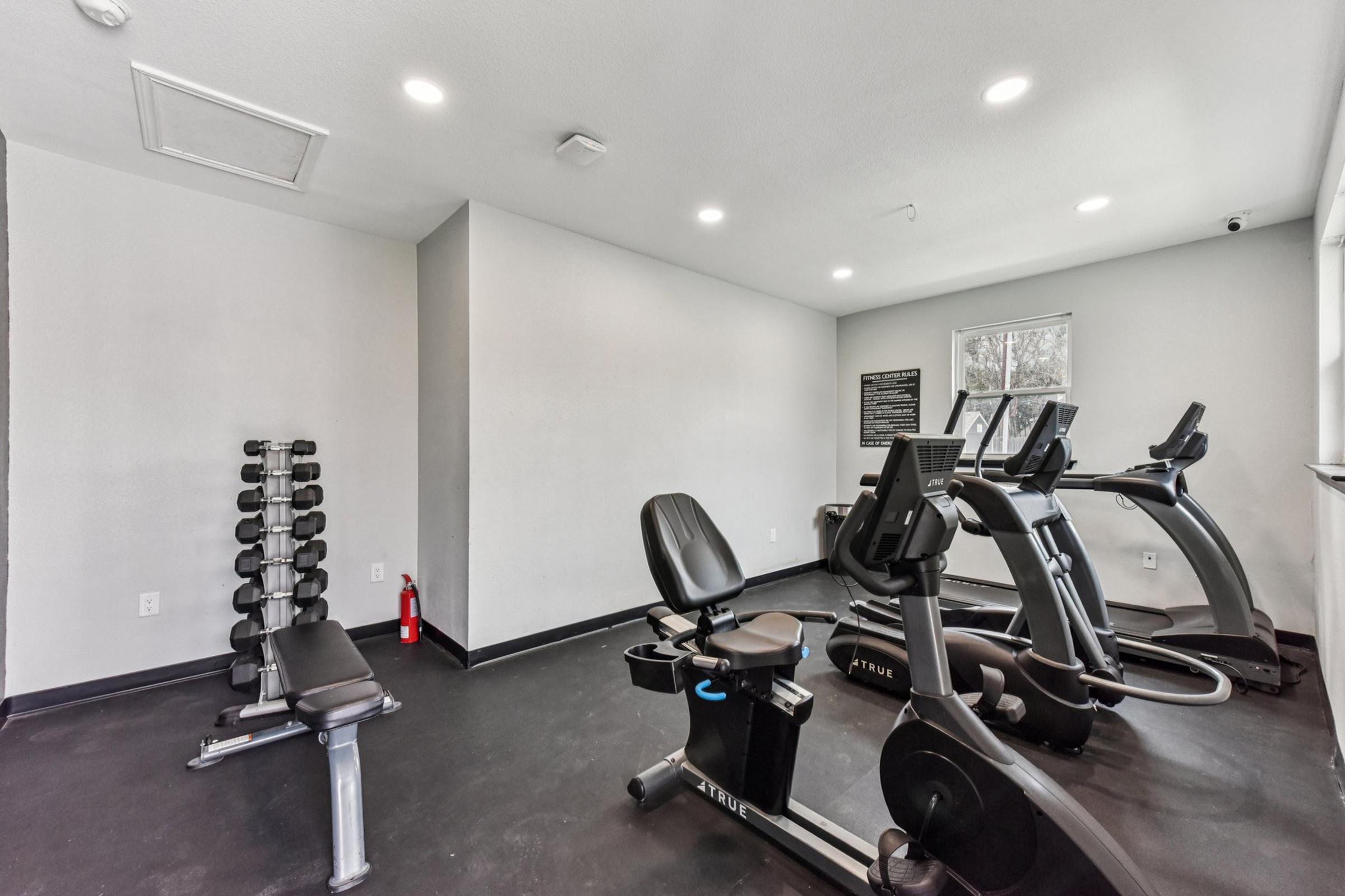 A modern, well-lit fitness room featuring a weight bench, a set of dumbbells, and two exercise machines. The walls are painted light gray, and there is a window letting in natural light. A fire extinguisher is visible in the corner.