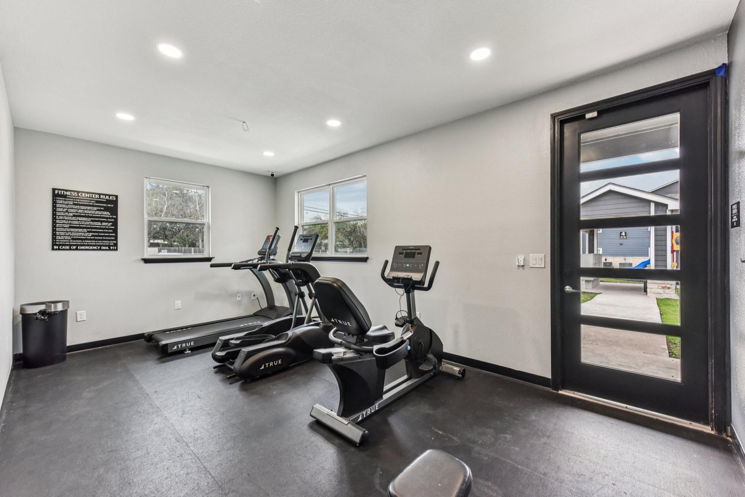 A modern fitness room with two elliptical machines and a treadmill. Large windows provide natural light, and a sign on the wall lists fitness guidelines. The floor is black and there is a trash can in the corner. A glass door leads outside to a view of a house and yard.