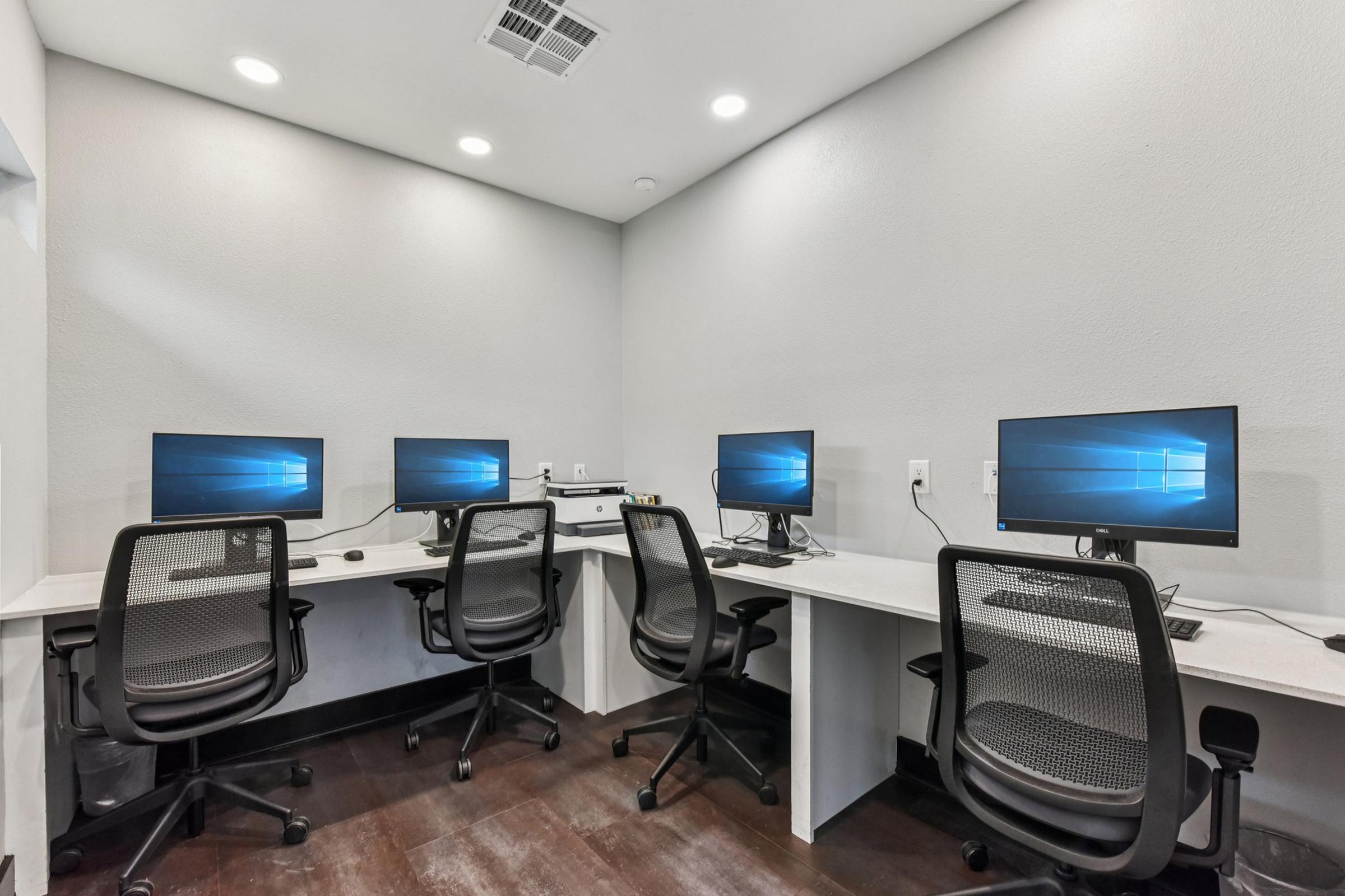A small, modern office space featuring four desks, each with a computer monitor, ergonomic black chairs, and a clean, minimalist design. The walls are painted a light color, and the flooring is dark. Soft overhead lighting illuminates the area, creating a professional atmosphere suitable for work or study.