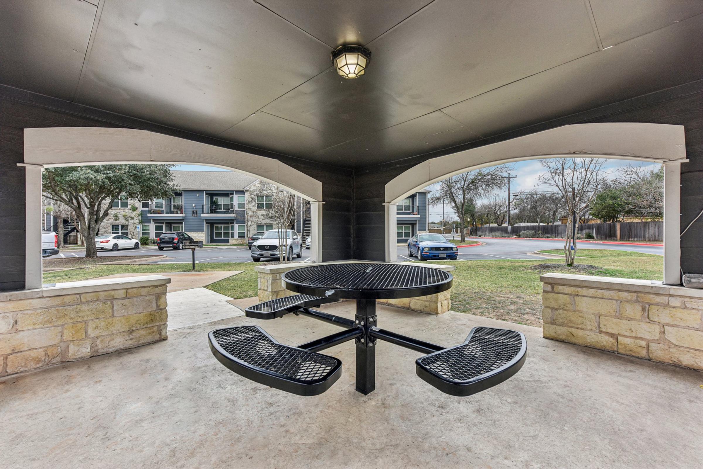 A covered outdoor seating area featuring a round black picnic table with attached benches, surrounded by stone walls. The view includes two windows showing parked cars and nearby buildings. The space is well-lit with a ceiling light, set in a residential environment with trees visible outside.