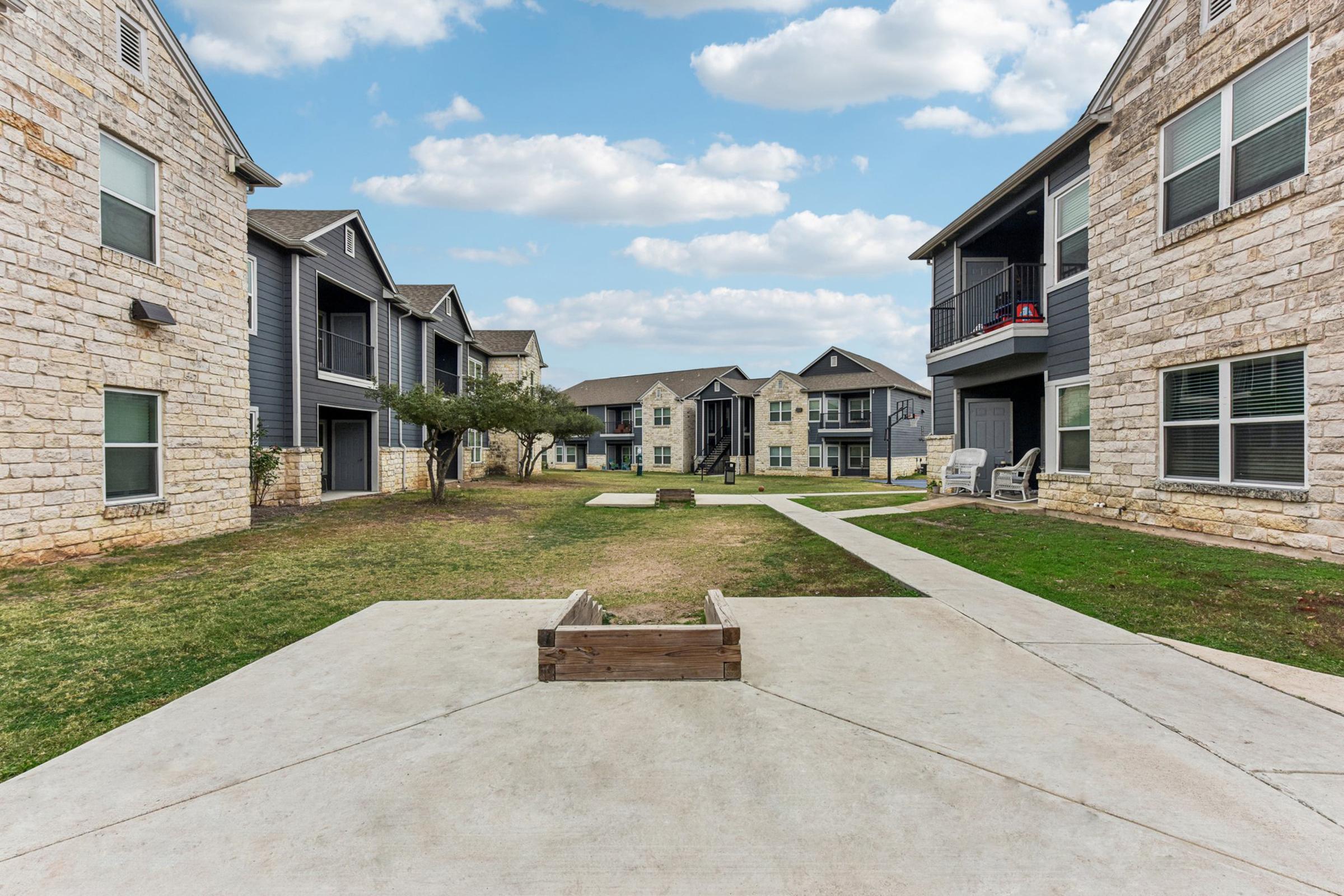 View of a residential complex featuring two-story stone and wooden buildings arranged around a grassy courtyard. A concrete pathway leads through the center, with a wooden structure on the ground. The sky is partly cloudy, enhancing the outdoor ambiance.