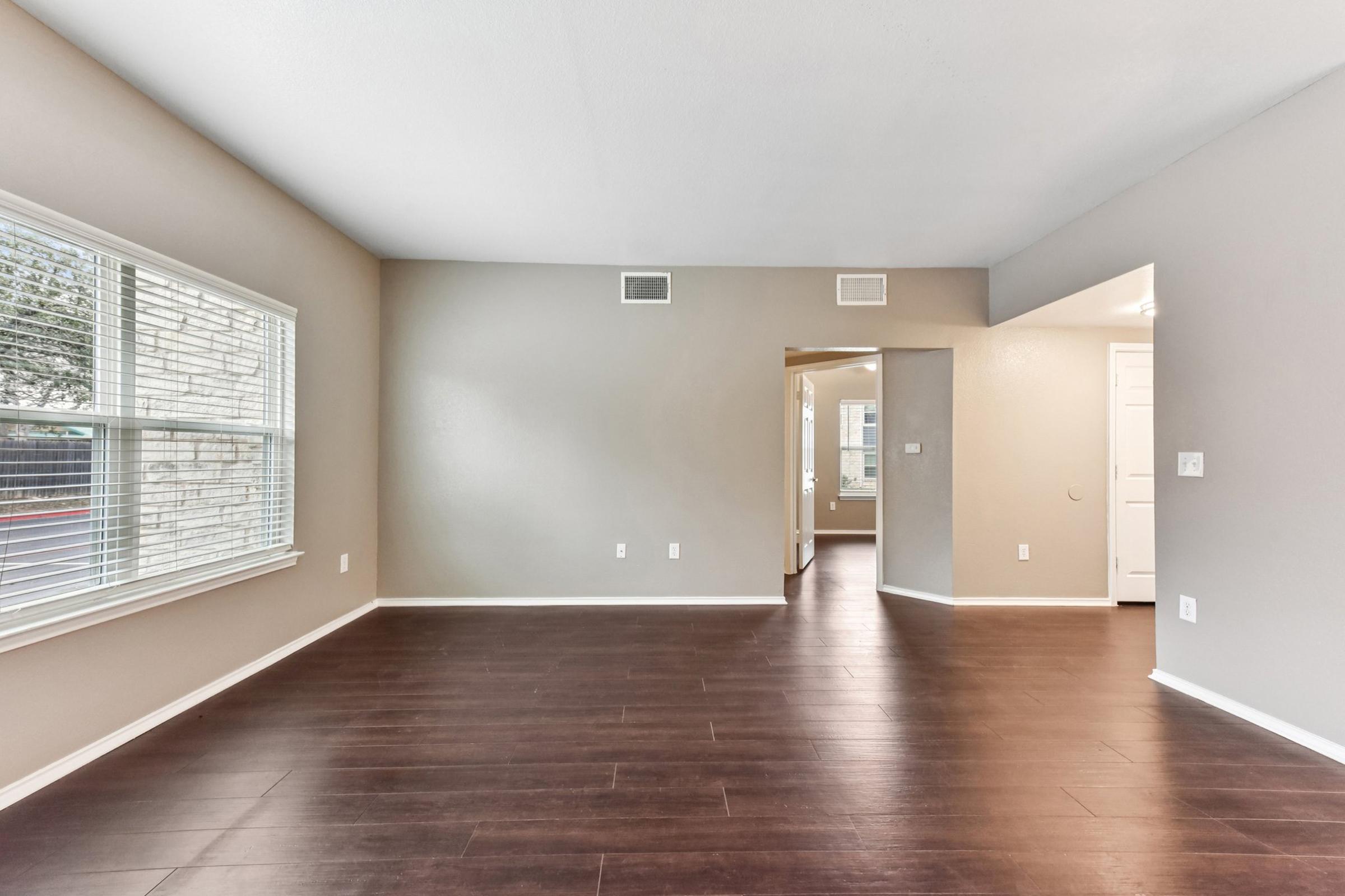 A spacious and bright living area featuring brown laminate flooring, light gray walls, and large windows that let in natural light. An open entrance leads to another room, and the overall design offers a clean and modern aesthetic, ideal for various décor styles.